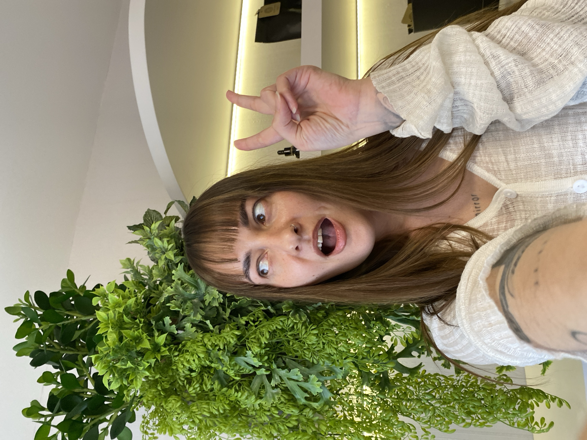 A young woman with brown hair and bangs makes a funny face at the camera, sticking out her tongue. She is surrounded by green foliage, possibly decorative plants. She wears a white blouse with ruffled sleeves. She has a tattoo on her neck that says "mirror" and tattoos on her left arm. She is making a gesture with her right hand with three fingers extended and joined at the tips. The background shows a white wall with recessed lighting and a dark object hanging on the wall. There is no discernible evidence in the image to indicate that the location is Votkinsk, Russia.