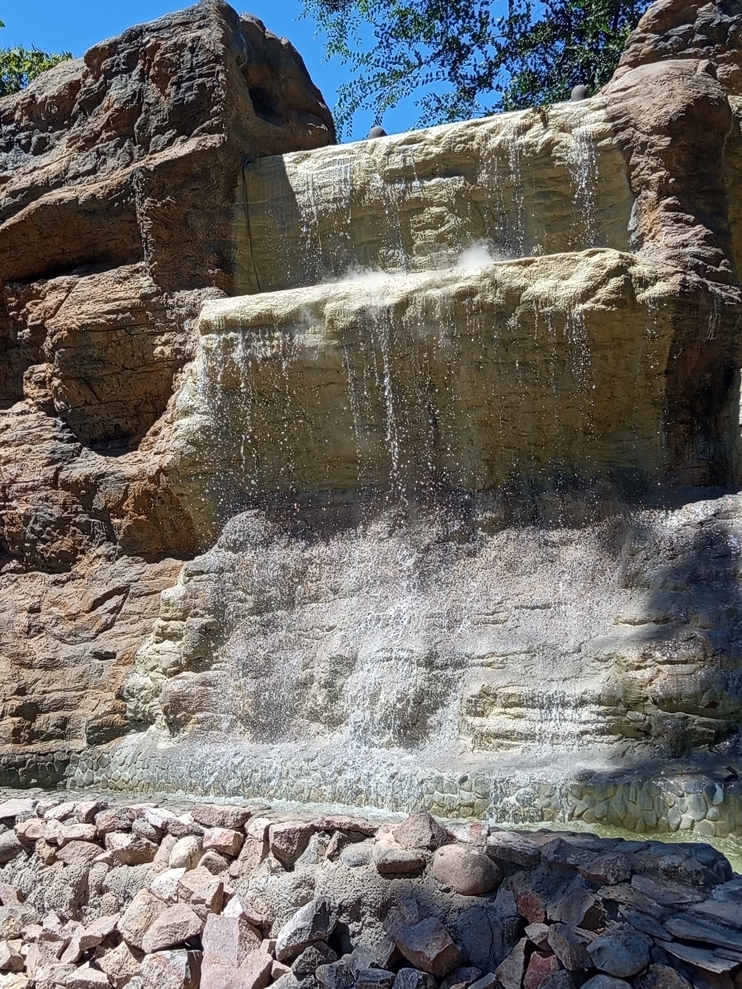 The image shows a man-made waterfall with a textured rock face, cascading down in multiple tiers. The water is falling onto a rocky base, with a pool of water at the bottom. The waterfall appears to be surrounded by a rocky landscape and some greenery is visible at the top of the image. The sky is blue and clear. This waterfall is located in the Botanical Garden of Shymkent, Kazakhstan.
