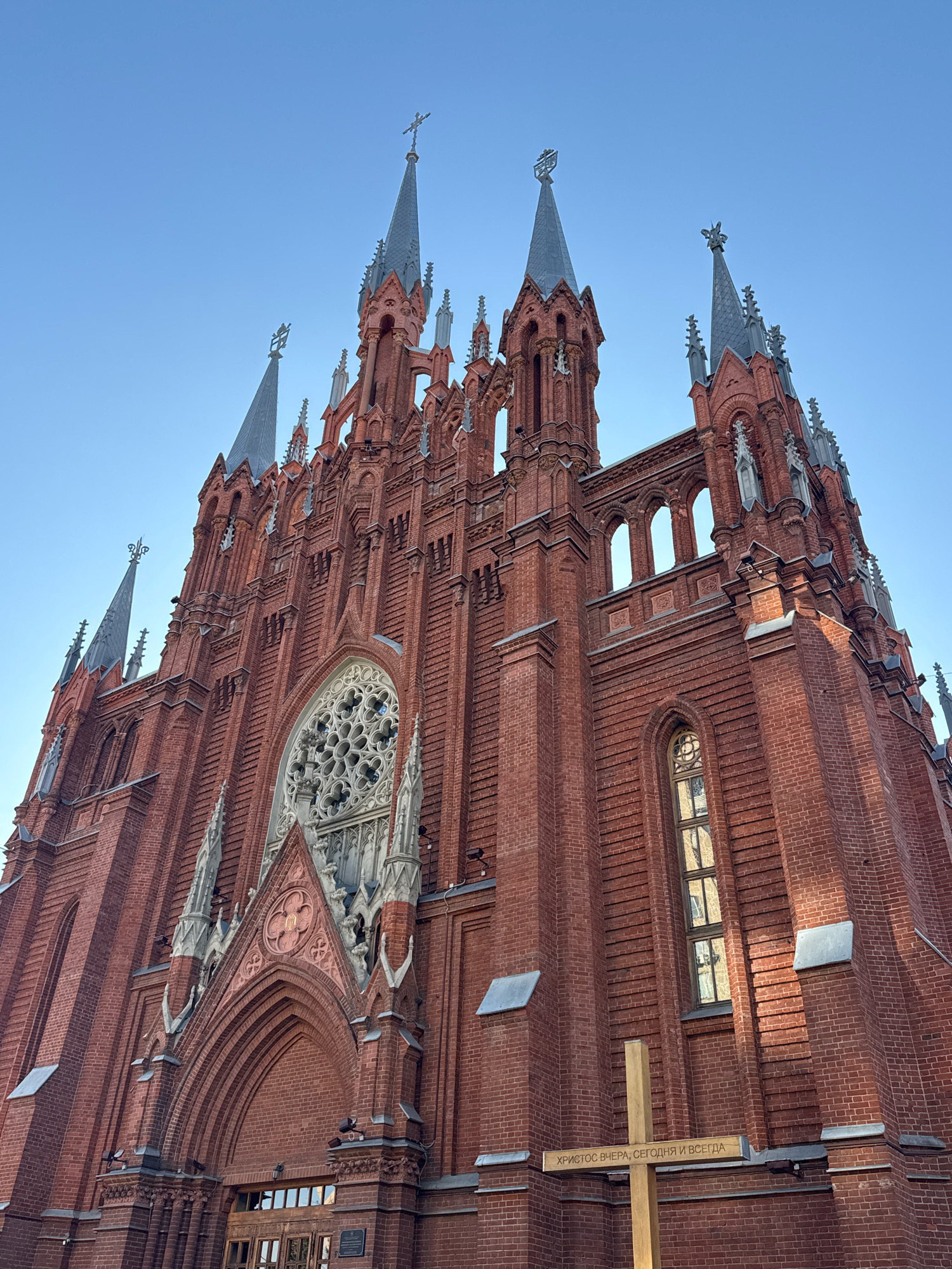 The image shows the Moscow Cathedral of the Immaculate Conception, a Roman Catholic cathedral located in Moscow, Russia. The photograph is taken from a low angle, looking up at the facade of the red brick building. The sky is a clear, light blue. The cathedral features Gothic architectural elements, including spires, pointed arches, and a rose window. There is a wooden cross in the foreground with Russian text on it, which reads "ХРИСТОС ВЧЕРА, СЕГОДНЯ И ВСЕГДА" (Christ yesterday, today, and forever). No people are visible in the image.