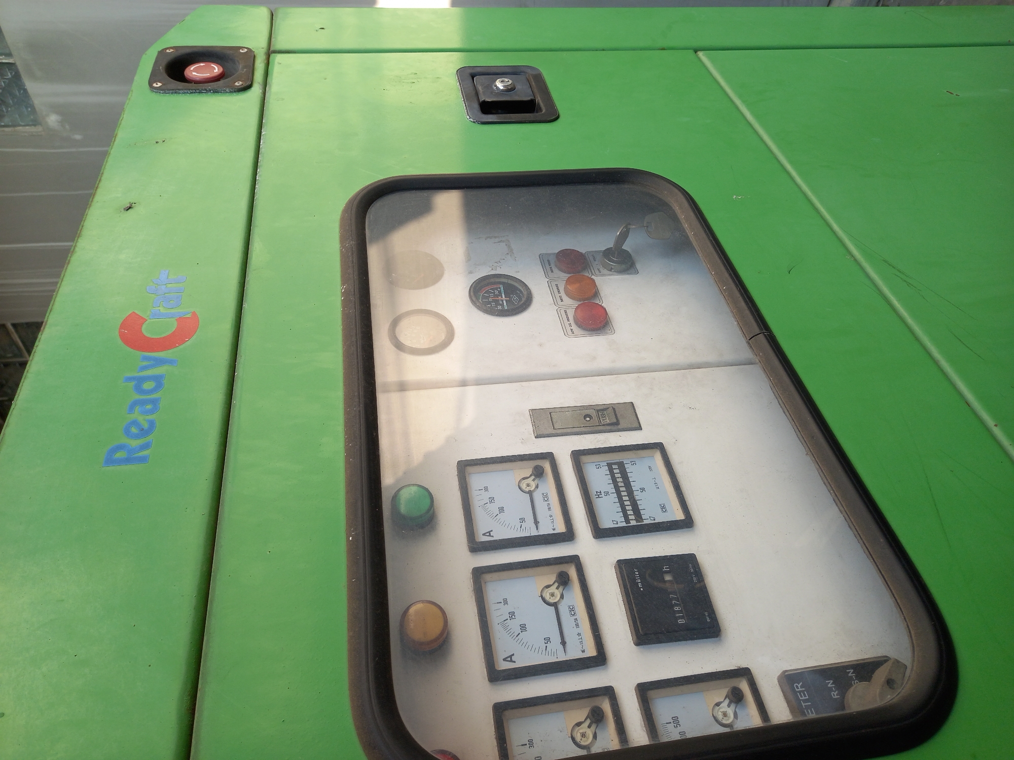 A green generator unit with a glass window is visible in the outdoor area of a city in Samarinda, Indonesia. The generator has a prominent "ReadyCraft" logo in blue and red on its upper section. The glass window reveals a control panel with various gauges and buttons. Some of the visible gauges include amperage meters, a frequency meter, and an hour meter displaying "01877". There are also several colored buttons, including red, orange, and green, and a key inserted into a switch labeled "CUT-OFF / STANDBY". A red emergency stop button with a twist-to-release mechanism is located on the upper right side of the generator. The surrounding environment is mostly out of focus but appears to be an industrial or service area.