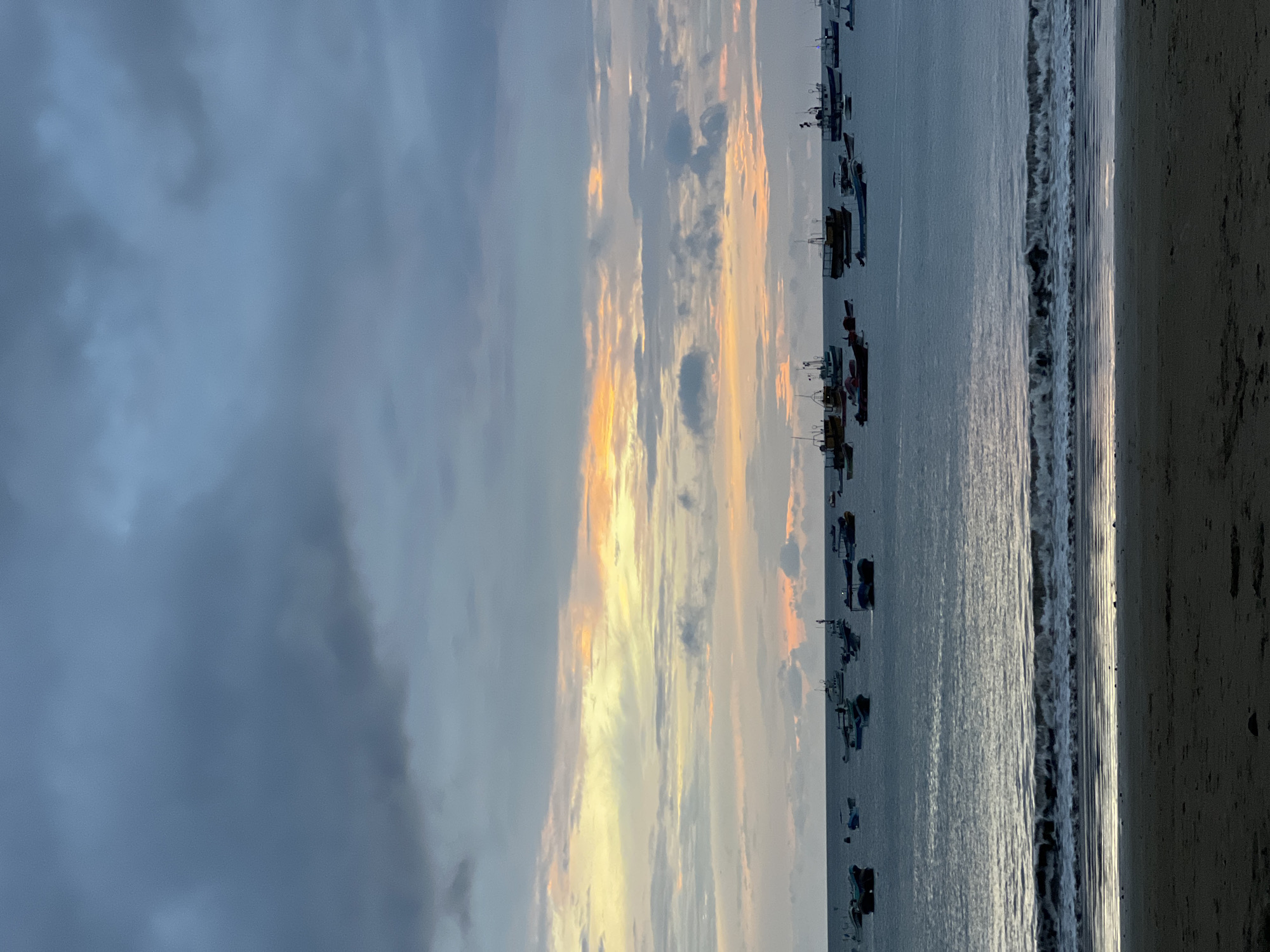 This image depicts a tranquil scene at sunset on the beach in San Juan del Sur, Nicaragua. The sky is a canvas of soft hues, with a blend of twilight blues, greys, and touches of orange and yellow from the setting sun. A gentle wave breaks on the sandy shore in the foreground, reflecting the muted colors of the sky. In the mid-ground, numerous fishing boats are anchored on the calm ocean, their silhouettes dark against the water. The horizon is a clear line separating the sea from the sky, with the clouds adding texture and depth to the atmospheric lighting. The overall impression is one of peaceful solitude and the quiet end of a day.