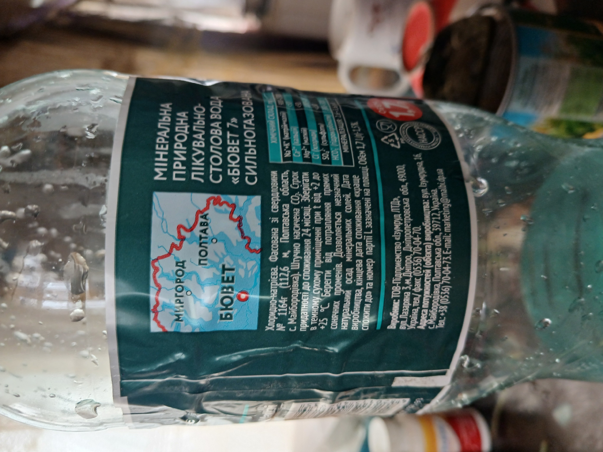 This is a close-up, eye-level, indoor shot of a partially filled plastic bottle of "BiuVet 7" mineral water, a Ukrainian brand. The bottle is covered in condensation. The label is predominantly teal and white, featuring a map of Ukraine with points marked as "Myrhorod" and "Poltava." Text on the label describes the water's properties, chemical composition, and manufacturer. The manufacturer's details indicate it is from the Dnipro region and also mentions an address in Poltava Oblast, specifically the town of Hlushky, Ukraine. To the right of the bottle, a white mug and a partially visible can are present on a surface, suggesting a domestic or casual setting. The lighting is natural and appears to come from the left, casting soft shadows. There are no people visible in the image.