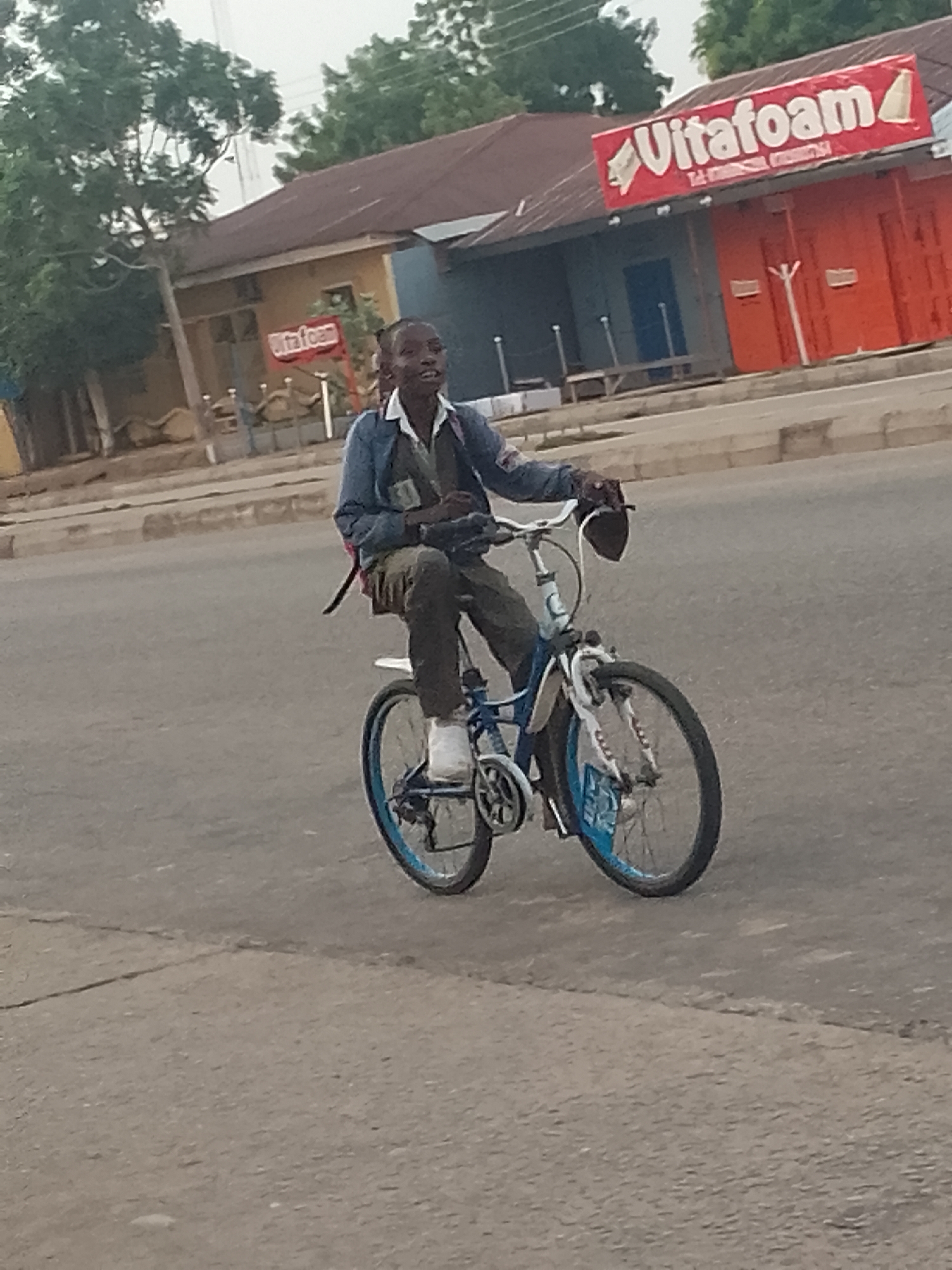 A young person, likely male, is riding a bicycle on a paved road in Pantame, Nigeria. The person is wearing a dark jacket over a white shirt and dark pants, with white sneakers. They are looking forward with a slight smile. The bicycle is blue and white with visible training wheels. In the background, there is a building with a red sign that reads "Vitafoam" and other text in smaller font. The building has a dark-colored roof and red-colored walls. Trees are visible behind the building. The road surface appears worn with some cracks. The sky is overcast.