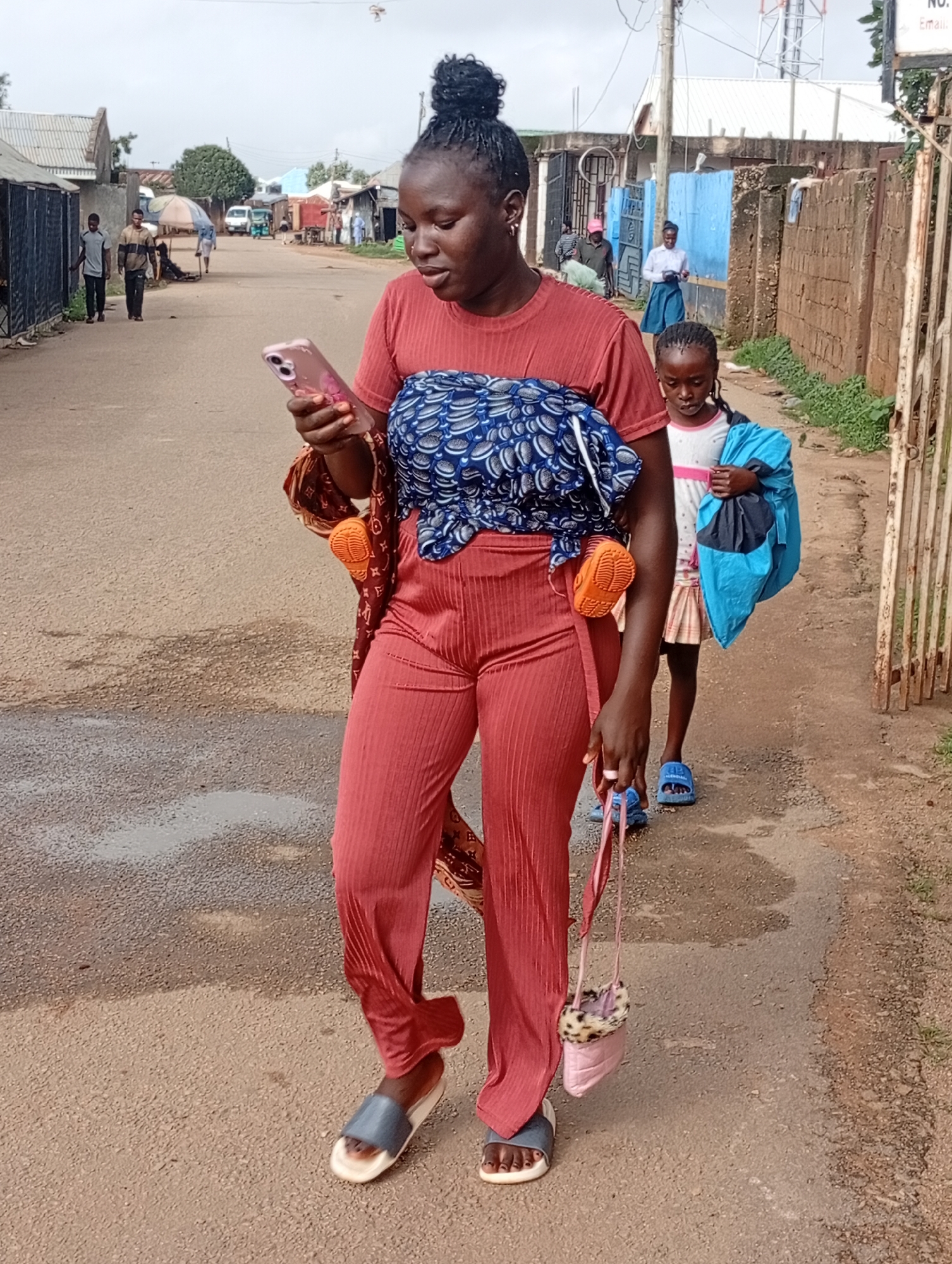 A woman in a red outfit and a child walk down a street. The woman is carrying a pink cell phone and wearing a blue and white patterned wrap around her chest. The child is carrying a blue bag. The street is paved with asphalt and lined with buildings. The sky is overcast.