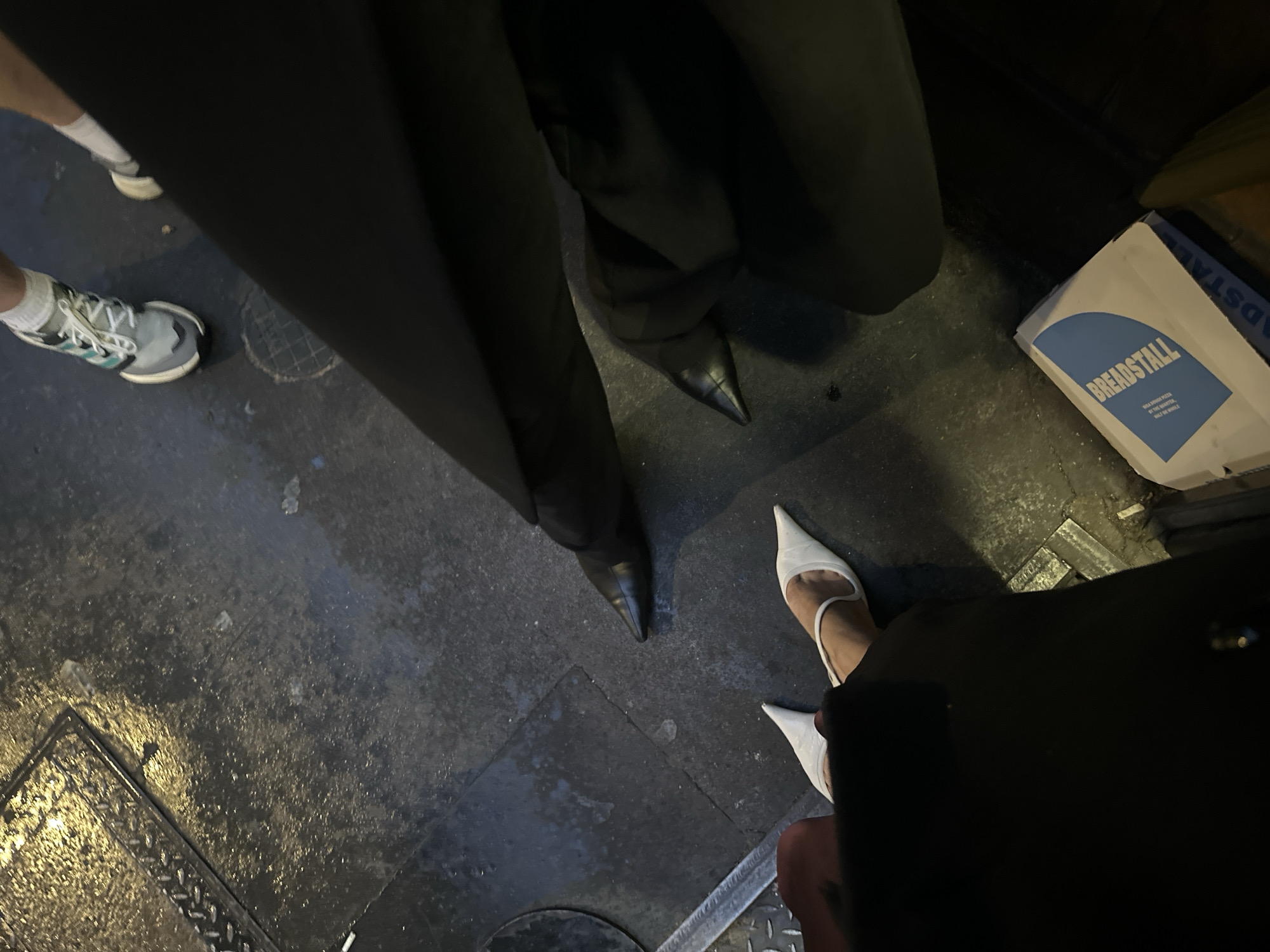 The image shows a view of several people's feet standing on a street in London. The person on the left is wearing grey and white sneakers with white socks. In the center, someone is wearing black dress shoes with black pants. Next to the black shoes, a person is wearing white pointy heels. There is a white box on the right side of the image labeled "Breadstall" with the tagline "Wild dough pizza at the Oyster, Half the Whale". The ground appears to be wet and there is a drain grate visible on the lower left of the image. The background and overall scene suggest an urban environment.