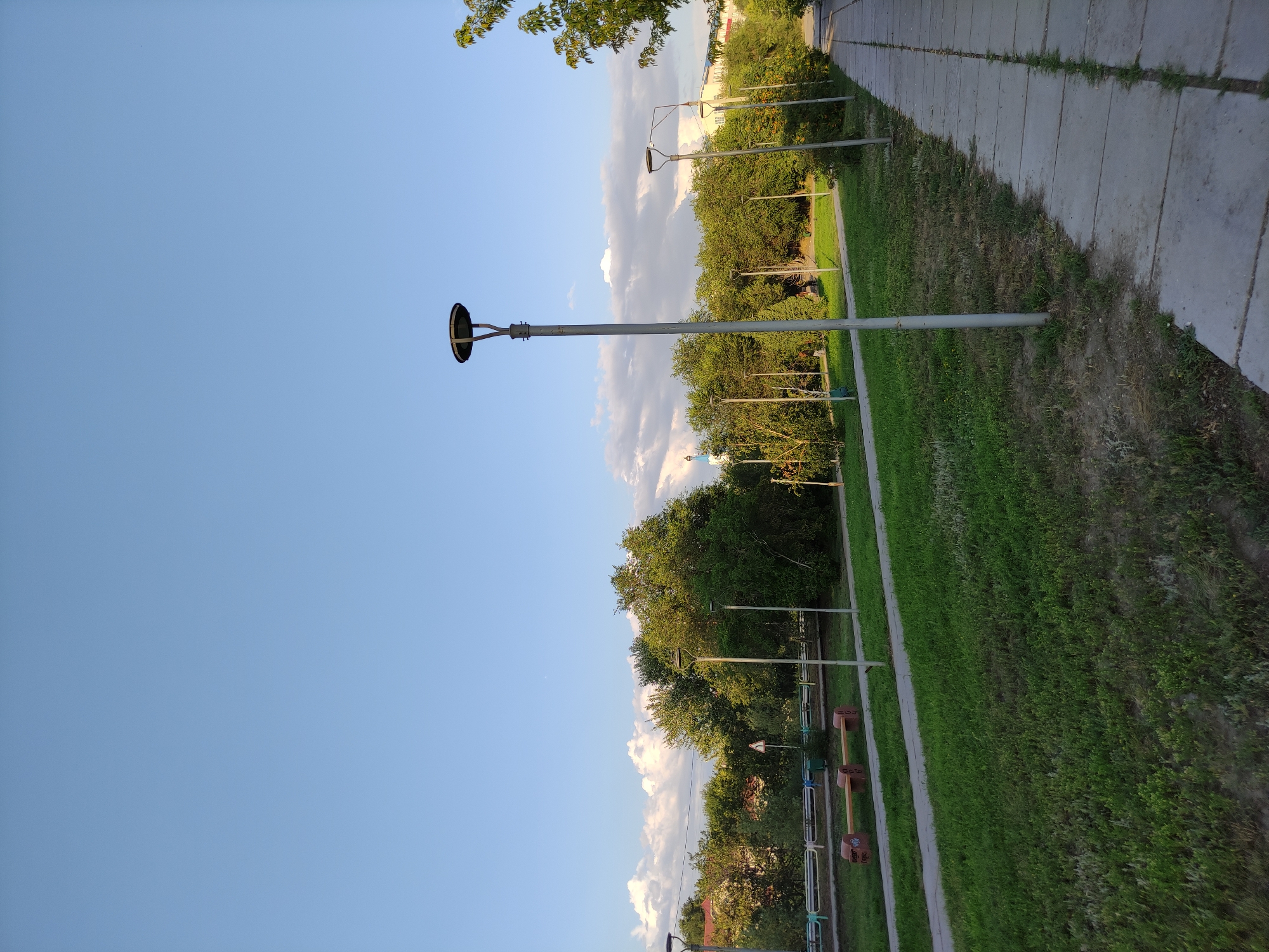The image depicts a paved pedestrian walkway adjacent to a grassy area with trees and park benches. The sky is clear with some scattered clouds. Several lampposts are visible along the walkway. In the background, there is dense foliage, buildings, and a white fence. The setting appears to be a park or a residential area in Semey, Kazakhstan. There are no people visible in the image. The overall impression is one of a serene, public outdoor space during daylight hours.