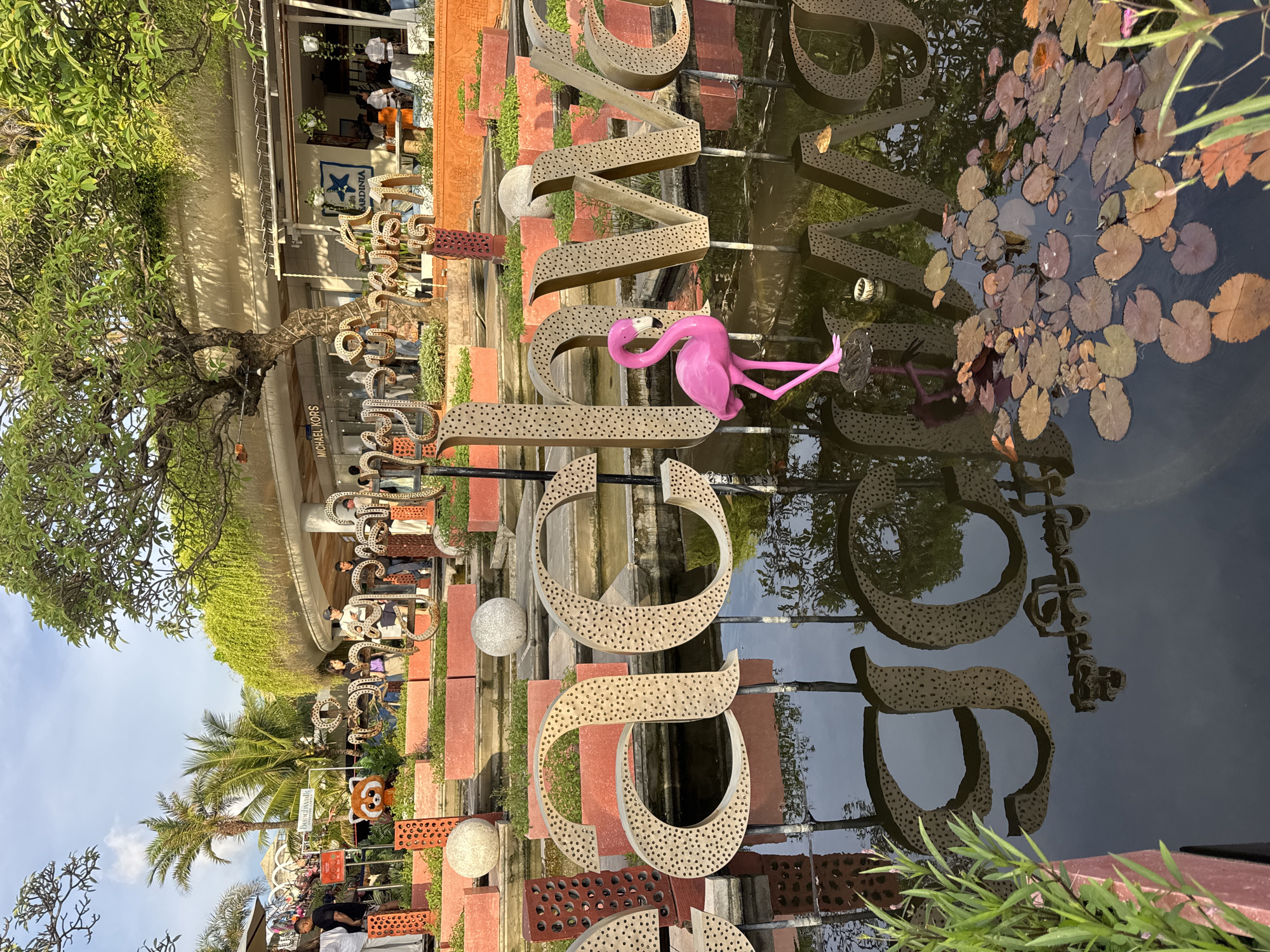 This image shows a decorative installation spelling out the word "LOVE" in Kuta, Indonesia. The letters are made of a perforated, light brown material and are positioned partially submerged in a pond with lily pads. A bright pink flamingo statue sits atop one of the letters. The letters are mounted on a series of red brick structures, with some vegetation interspersed.
In the background, there are trees, buildings, and people walking around. Some shops and signs can be seen, including one that says "Michael Kors" and another with "Cafe Sardinia". The overall atmosphere is sunny and outdoorsy, suggesting a tourist area or public space. The reflection of the installation and surrounding environment is visible in the pond's surface.