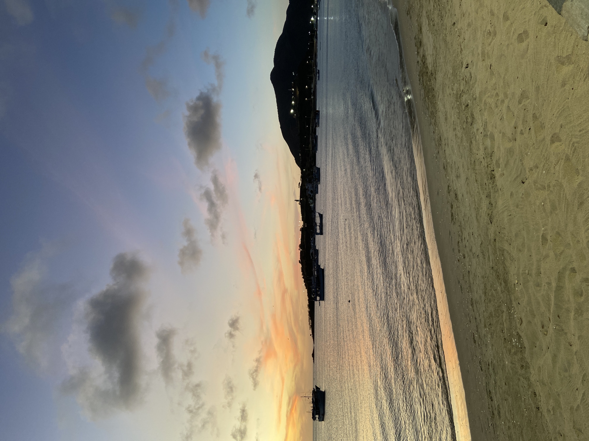 This image presents a serene coastal landscape at twilight, likely sunset, in Juan Griego, Venezuela. The scene is composed of a vibrant sky, a calm body of water, a mountainous coastline, and a sandy beach.
The upper section of the frame is dominated by a beautifully colored sky, exhibiting a gradient from soft blues and purples in the upper left to warm oranges, pinks, and yellows closer to the horizon. Dark grey, scattered clouds are visible, contrasting with the luminous background and adding texture to the sky.
Below the horizon, a prominent mountain range or large hill creates a dark silhouette, stretching across the middle left of the image. At its base, numerous small, bright lights glow, indicating a town or settlement nestled along the shore. The calm waters of a bay or sea extend from the base of the mountain towards the right foreground, reflecting the warm, diffused light of the twilight sky. Several boats, appearing as dark shapes, are gently moored or floating on the water's surface.
To the right, a wide stretch of light-colored sand forms a tranquil beach, extending from the foreground to meet the water's edge. The sand displays subtle patterns and textures, possibly from gentle waves or footprints. The overall atmosphere is peaceful and picturesque, capturing the beauty of a coastal evening as day transitions to night. No people or explicit text are visible in the scene.