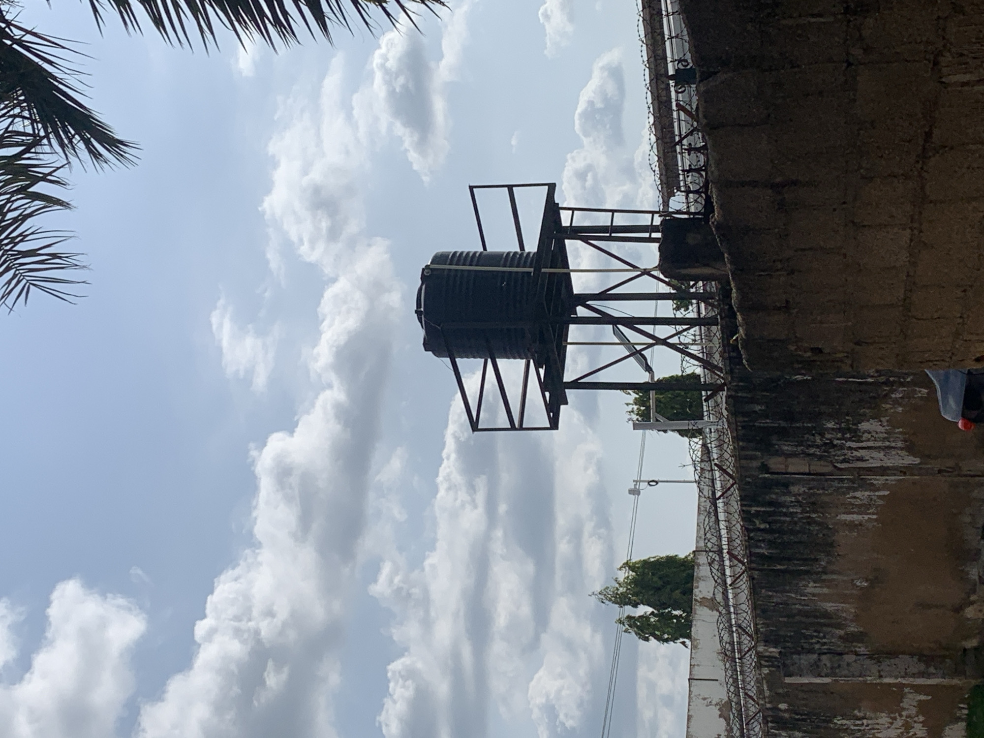 An outdoor, eye-level, medium shot shows a black water tank on a metal support structure. The tank is cylindrical with a ribbed exterior and sits atop a dark metal framework with crossbeams and a platform. The structure is positioned on the right side of the image, partially obscuring a white building in the background. A section of a stone wall with patches of discoloration is in the foreground, topped with barbed wire. Behind the wall, several tall, green trees are visible, along with power lines and a street light. The sky is a light blue with scattered white clouds. A portion of a palm frond is visible in the upper right corner of the frame. The overall scene suggests an urban or industrial setting with a focus on infrastructure.