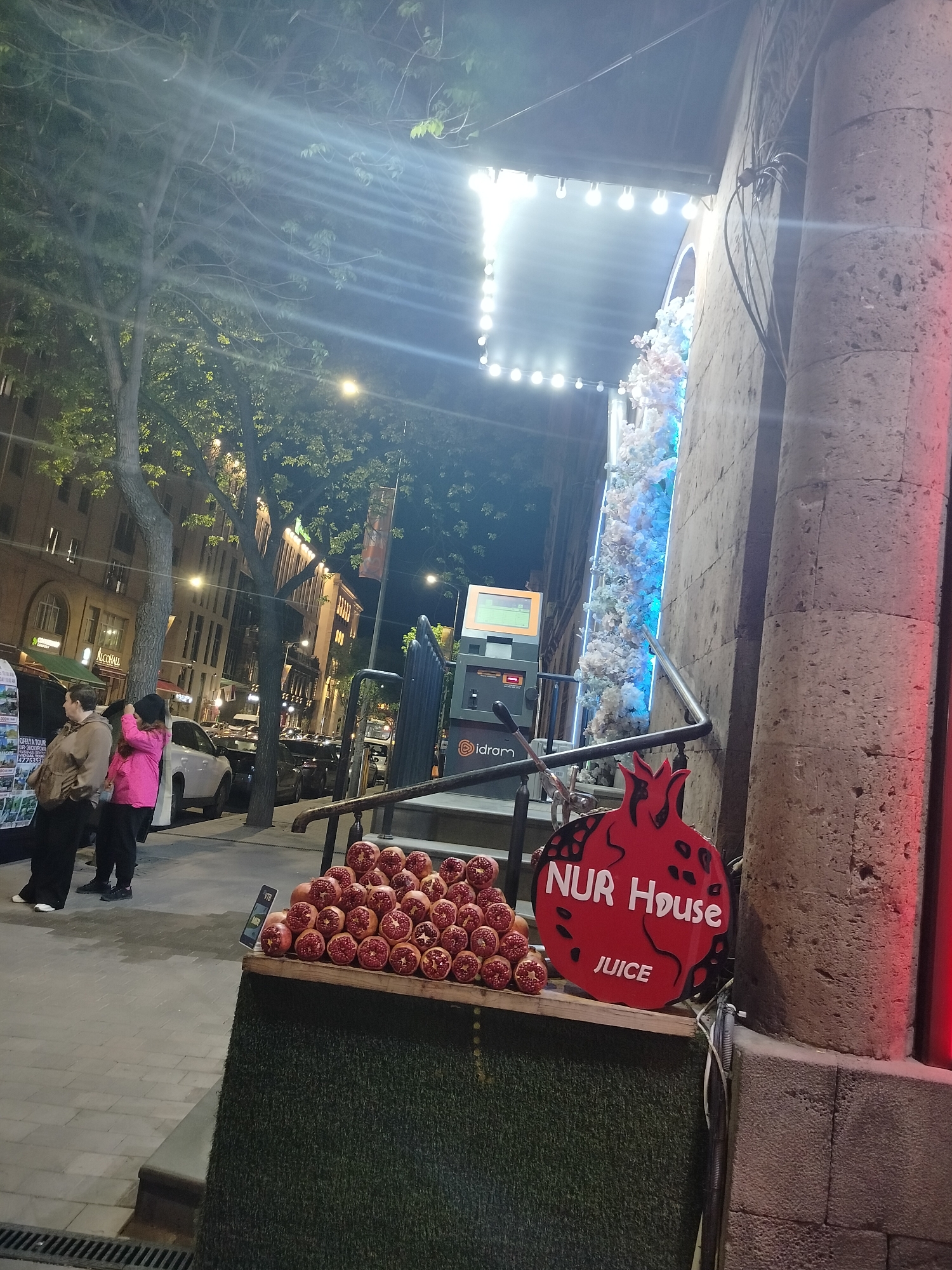 The image captures a street scene at night in Yerevan, Armenia. On the right is a building adorned with white floral decorations illuminated by blue lights. Adjacent to the building is a sign shaped like a pomegranate, advertising "NUR House JUICE." In front of the sign is a display of pomegranates. To the left of the display, two women stand on the sidewalk; one wears a pink jacket and black pants, while the other is in a tan jacket and black pants. In the background, a street lined with buildings stretches into the distance, with streetlights and cars adding to the urban setting. An "idram" payment terminal is also visible.