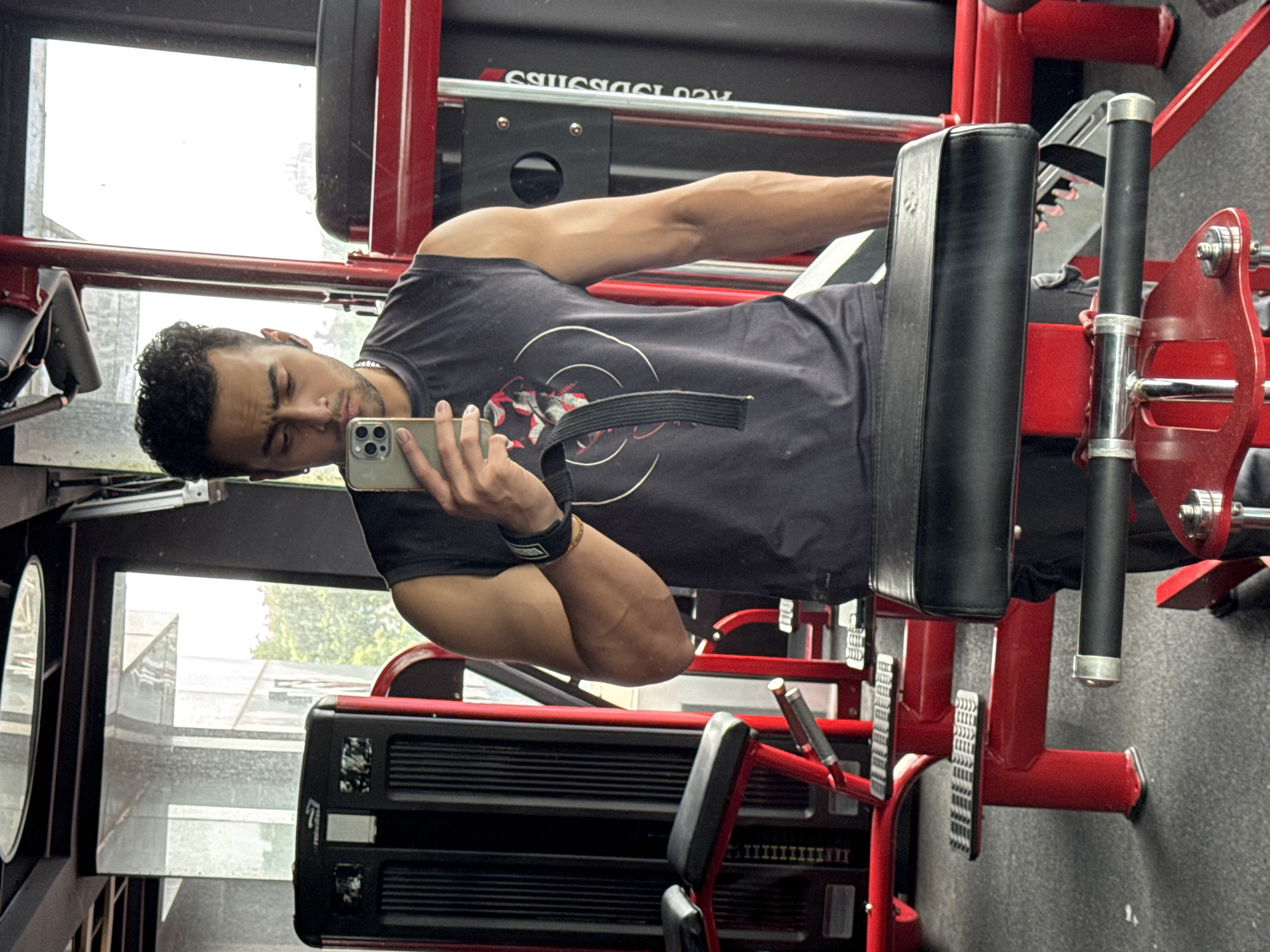 A man is taking a mirror selfie in a gym. He is wearing a sleeveless dark grey t-shirt and black pants. He has curly dark hair and is holding a smartphone in his right hand, which is raised to take the picture. He has a weightlifting belt around his waist and is wearing a wrist wrap on his left wrist. The gym equipment, including weight machines and benches, is predominantly red. The floor is grey and textured. Large windows with reflections of trees and buildings are visible in the background, suggesting an urban environment. The lighting is typical of an indoor gym. The location is identified as a gym in Kolkata, India.