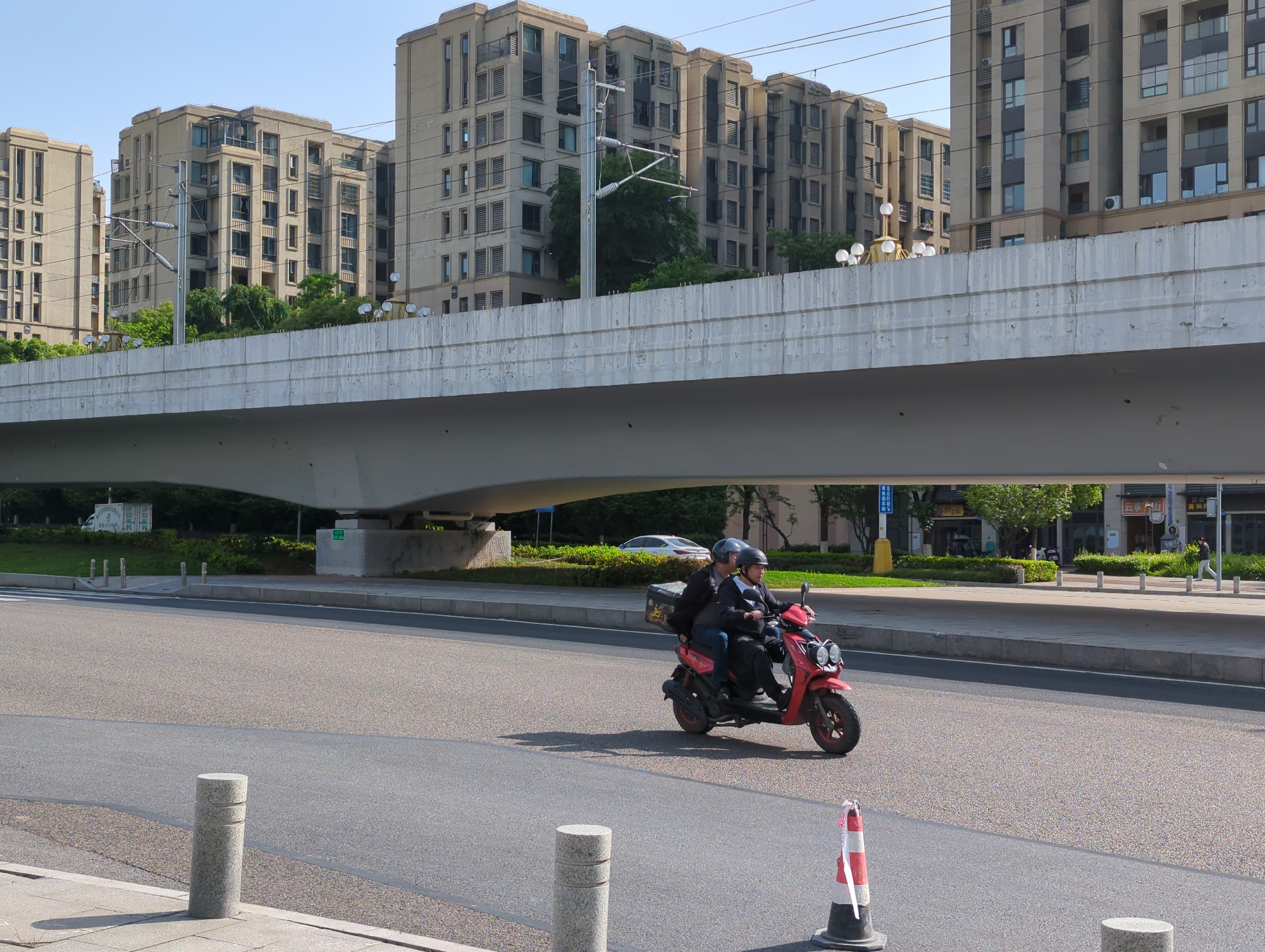 This image depicts a sunny urban street scene in Jiangjin, China, dominated by a large, elevated concrete overpass that stretches across the middle ground. Behind and above the overpass, several modern, multi-story residential buildings with numerous windows and balconies rise towards a clear blue sky. Green trees and bushes are interspersed throughout the landscape, appearing both behind the buildings and underneath the overpass.
In the foreground, two individuals are riding a bright red scooter on a paved road, moving from left to right. Both riders wear helmets and dark jackets. The person in the back has a large, dark box attached to the rear of the scooter, suggesting a delivery service. The strong sunlight casts clear shadows from the scooter and from cylindrical gray bollards lining the adjacent sidewalk, indicating it is likely midday. A red and white traffic cone with a white ribbon stands near the right edge of the road.
Under the overpass, a white car is partially visible, along with various signs and road infrastructure. A utility pole with complex wiring is seen between the apartment buildings and the overpass. In the far background, on a sign to the right, Chinese characters "云峰街" (Yunfeng Street) and "黄周" are discernible, providing specific location cues within Jiangjin. The scene captures a typical, active urban environment under bright, clear weather conditions.