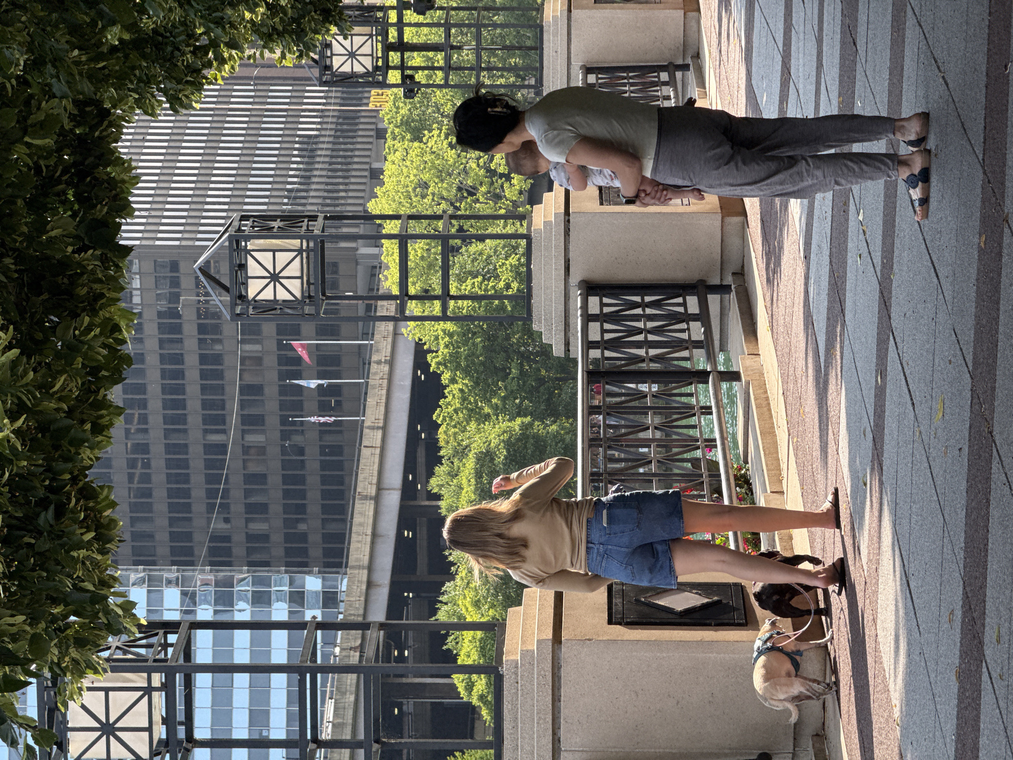 Two women and two dogs are on a paved walkway with a decorative metal railing. The woman on the left is facing away from the camera, wearing a tan long-sleeved shirt and denim shorts, and is holding the leashes of two dogs, a light-colored dog with a blue harness and a dark-colored dog. The woman on the right is also facing away, holding a baby, and wearing a light green t-shirt and gray pants. In the background, there are tall buildings and lush green trees. The scene appears to be along the Chicago Riverwalk in Chicago, Illinois.