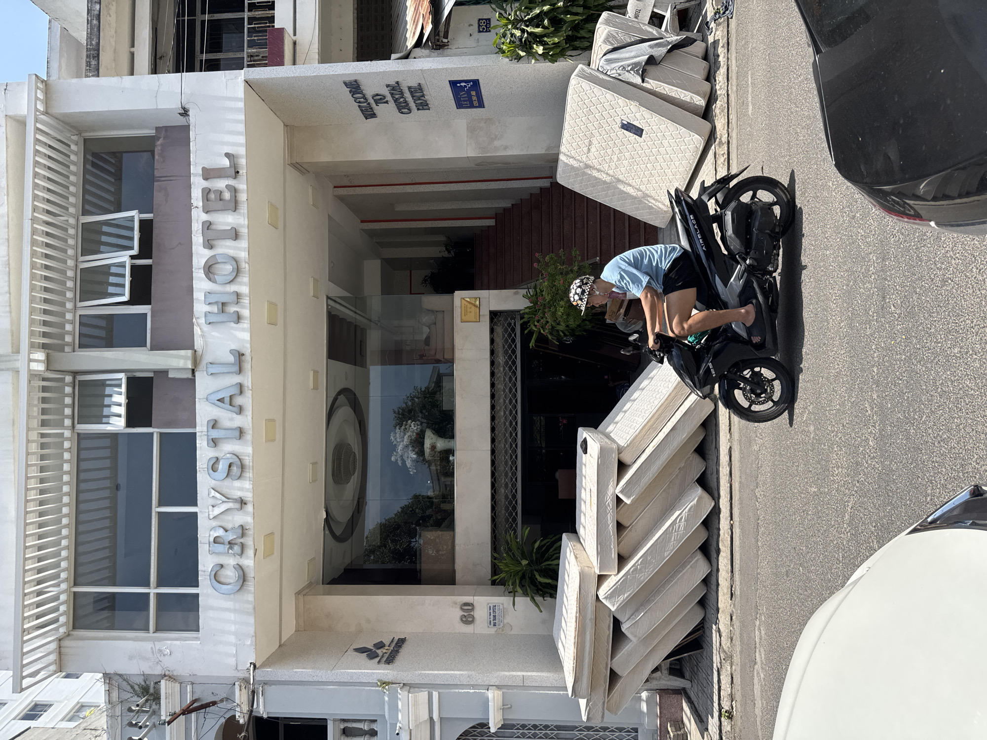 The image shows the entrance of the Crystal Hotel in Nha Trang, Vietnam, located at 60 Tran Phu street. A pile of mattresses is stacked against the hotel's facade, some leaning against the wall and others propped up by wooden supports. A person is riding a black scooter in front of the hotel, passing by the mattresses. The hotel has a large glass entrance and a set of stairs leading up into the building. The street in front of the hotel is paved and appears to be a busy area with other vehicles visible. The sky is clear and the weather appears to be sunny. The overall scene suggests a possible renovation or removal of old mattresses from the hotel.