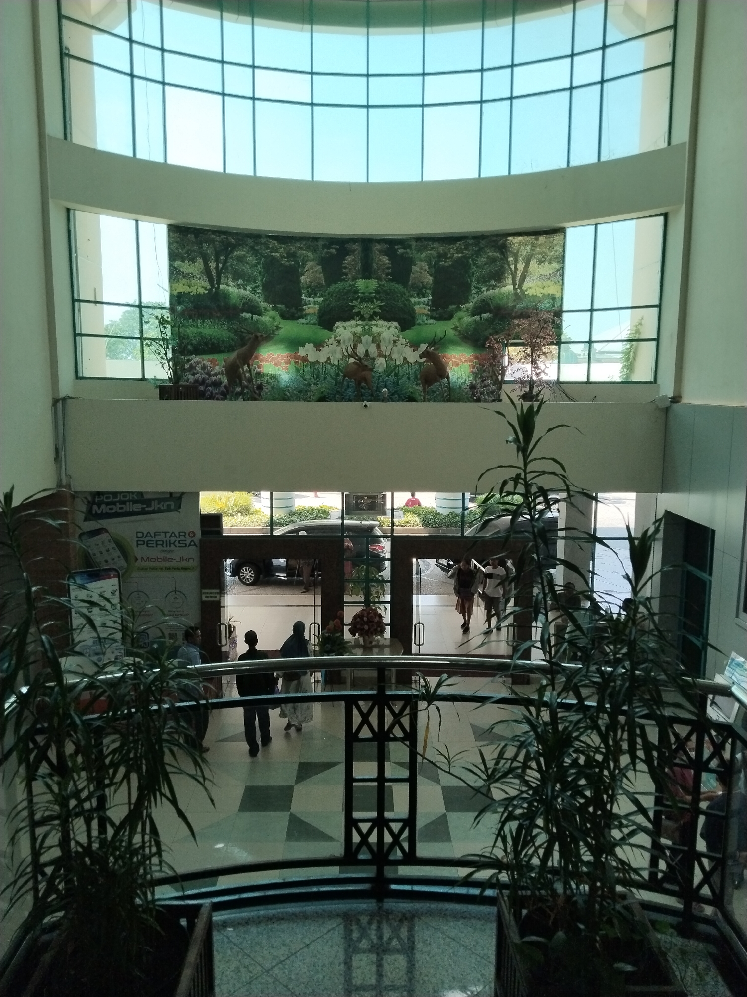 The image captures an interior view of a building, likely a public space, with an atrium and multiple levels. In the foreground, large potted plants with thin, dark green leaves are visible, obscuring parts of the scene. A dark metal railing separates the viewer from the lower levels.
The central part of the image showcases an open atrium with a checkered tile floor. Several people are walking on this level. To the left, a large banner or poster is displayed, featuring text and graphics, including a smartphone illustration, and the words "POJOK Mobile-Jkn DAFTAR Mobile-Jkn". Further back, an open doorway leads to an exterior area where a dark-colored car is parked.
Above the atrium, a wide decorative mural is mounted on the wall, depicting a lush garden scene with trees, flowers, and two deer. The mural is framed by a large, curved window with a grid pattern of blue-tinted glass. This upper window section is part of the building's architecture, offering a view of the sky or an upper exterior area. The building's interior walls are painted a light, neutral color. The overall impression is of a modern, well-lit interior space with decorative elements.
The location of Margorejo, Indonesia, is mentioned in the prompt but is not visually evident from the provided image alone. The image depicts an indoor scene typical of a hospital, a large office building, or a public service center, possibly in a region with such facilities.