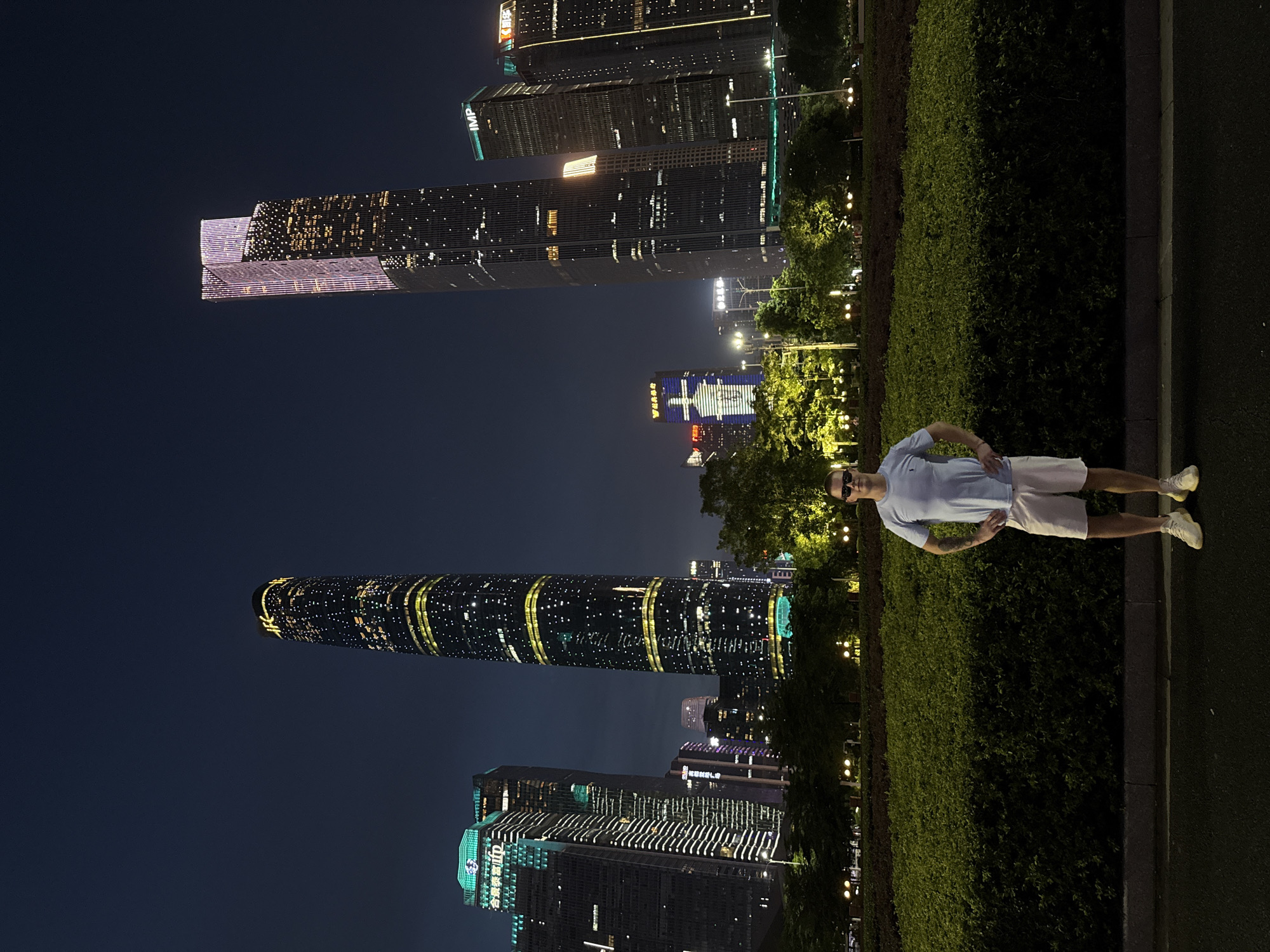 The image captures a nighttime scene in Tianhe District, China, featuring a man standing in the foreground. He is wearing a light blue t-shirt, white shorts, and white sneakers, with sunglasses on. His hands are placed on his hips, and he is looking towards the camera.
Behind him, a lush green hedge forms a boundary, and beyond that, a cityscape of illuminated skyscrapers dominates the background. The buildings are tall and modern, with various lights displaying patterns and outlines. One distinctive cylindrical building is lit with yellow bands. Another building displays a large blue and white graphic. The overall scene is characterized by the bright lights of the urban environment against the dark night sky, suggesting it is after sunset. The man appears relaxed, posing for the photograph. Visible text on the buildings includes "IMP" on one of the skyscrapers and Chinese characters on others. The presence of streetlights and illuminated trees adds to the vibrant city atmosphere.