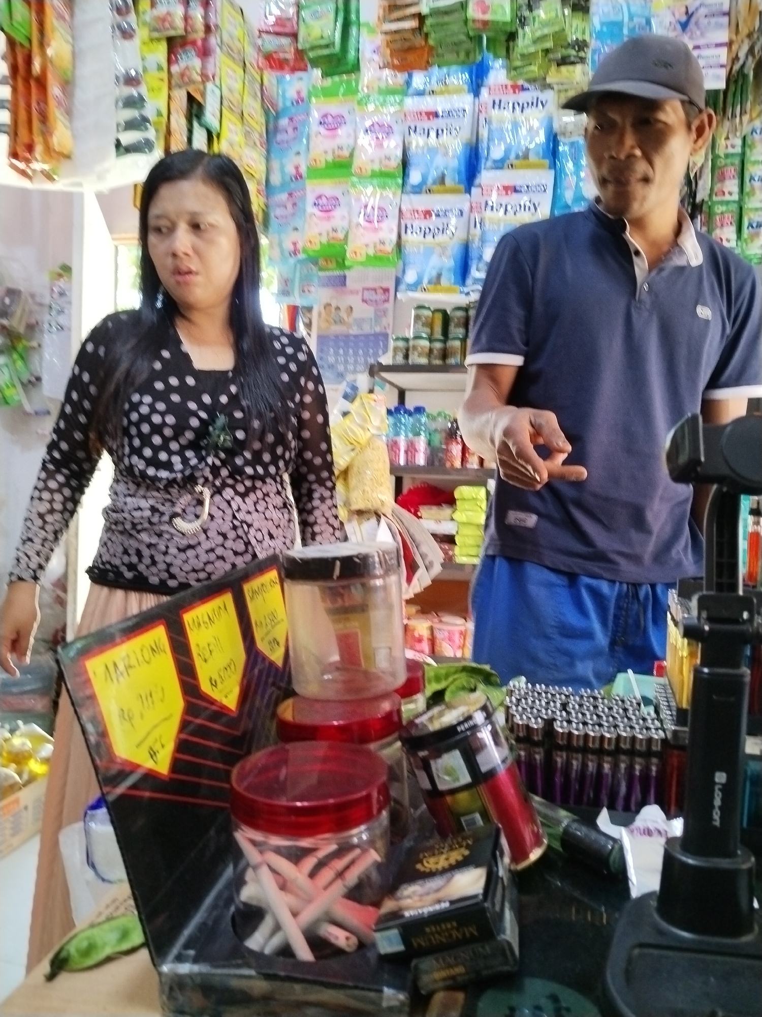 Two individuals, a woman and a man, are standing in what appears to be a small convenience store in Sawit Seberang, Indonesia. The woman, on the left, is wearing a black and white polka dot top and a light-colored skirt. She has dark hair and is looking to her left with a neutral expression. The man, on the right, is wearing a dark blue polo shirt and blue shorts, with a gray baseball cap. He is looking towards the camera and has his right hand extended, with his index and middle fingers making a gesture.
The store is filled with various products displayed on shelves and in containers. Among the visible items are packaged snacks and beverages, plastic containers with what appear to be cigarettes, lighters, and other small consumer goods. A yellow price tag with writing in Indonesian is visible in the foreground. The overall lighting suggests an indoor environment, possibly with natural light entering from the side. The scene depicts a typical retail setting in a local Indonesian market.