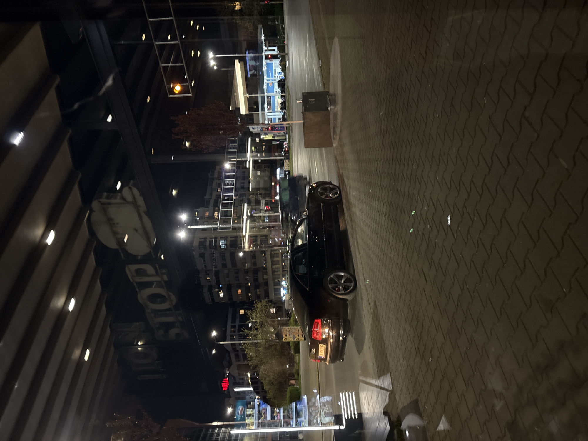 The image depicts an urban scene at night in an unknown city. A black car is parked along the curb on a paved street. A multi-story building with illuminated windows and signage is visible on the left side of the image. Streetlights and other buildings are visible in the background. A bus stop appears to be located nearby. The overall scene is illuminated by artificial lighting.