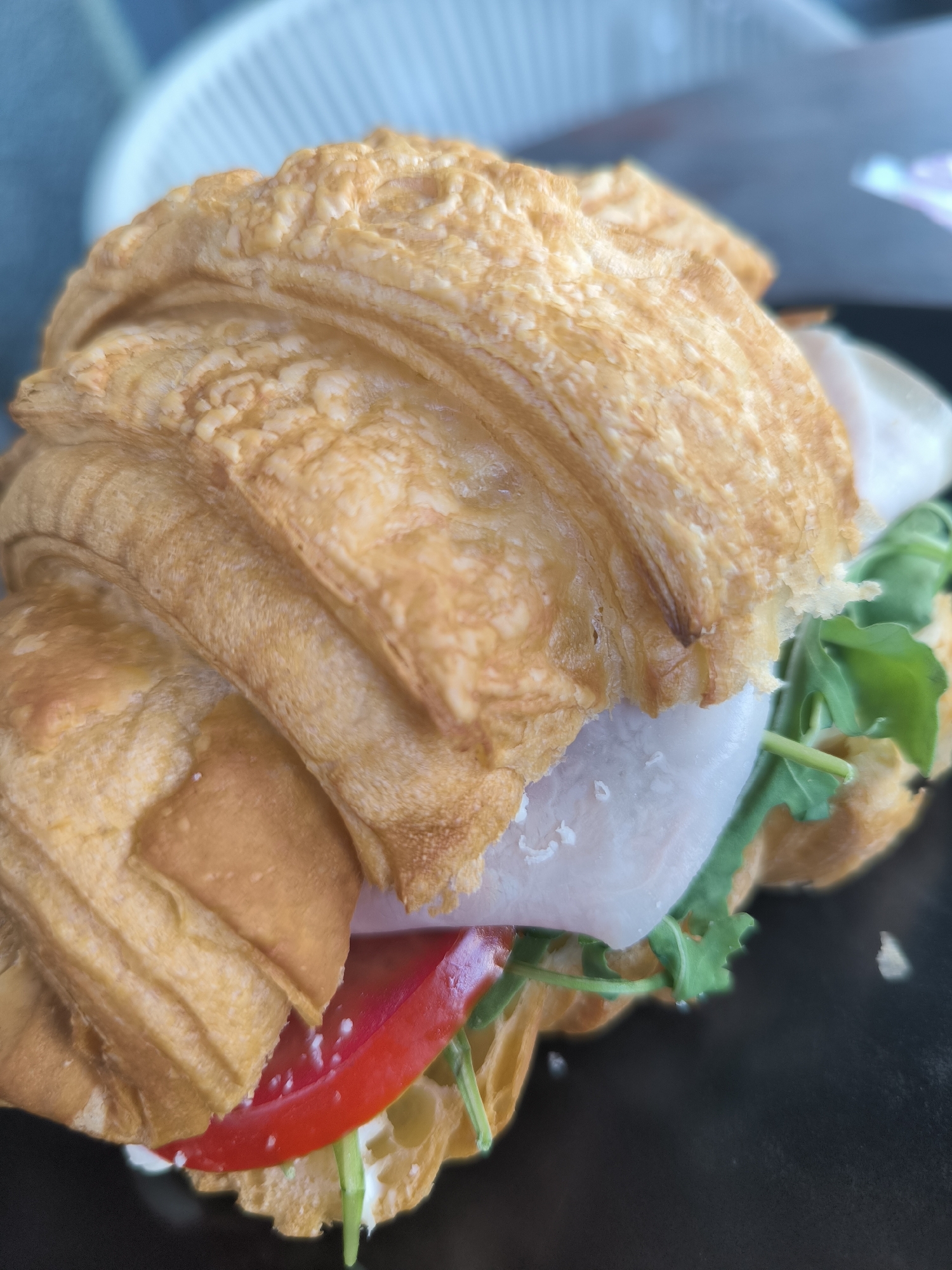 A close-up shot captures a ham and tomato croissant sandwich on a black plate. The croissant, golden brown and flaky, is filled with thinly sliced ham, a bright red slice of tomato, and fresh green arugula. The background is blurred, showing a light blue and white striped object, possibly part of a chair or table setting. The image does not provide any information about the location in Chisinau, Moldova.