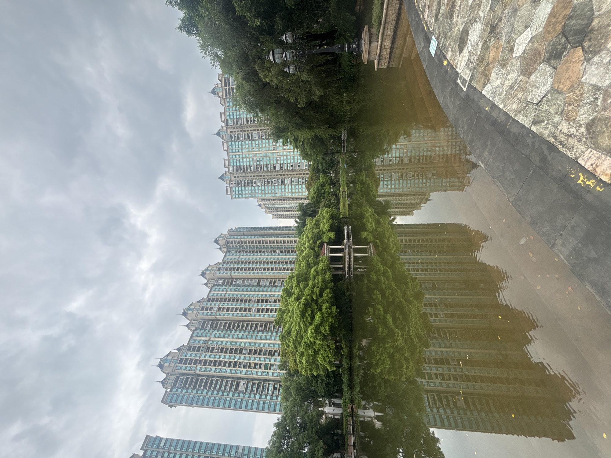 This is a full-shot, eye-level, outdoor, natural-light photograph of a tranquil park scene. The sky is overcast with visible clouds. In the background, there are several tall, modern apartment buildings with light-colored facades and turquoise windows. These buildings reflect in the murky water of a pond in the foreground. The pond is bordered by lush green trees and a small, ornate pavilion with white columns and a red roof. A black, multi-headed lamppost stands on the right side of the frame, situated on a stone pathway. The pathway is made of irregularly shaped stones in earthy tones. The image captures a sense of calm and urban nature, likely in a residential area within Nanhai District, China. There are no people visible in the photograph.