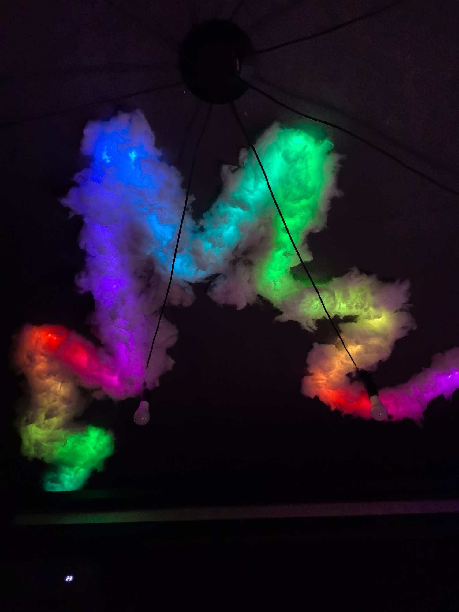 The image shows a darkened interior space with a ceiling fixture holding illuminated cotton-like formations. The formations glow in a rainbow of colors, including blue, green, purple, red, and yellow. Thin black wires suspend from the fixture and run through the "clouds," with a single light bulb hanging from one of them. The bottom of the frame shows a dark surface with a faint horizontal line. No people are visible. There is no indication this photo was taken in the city of Noviy, Russia.