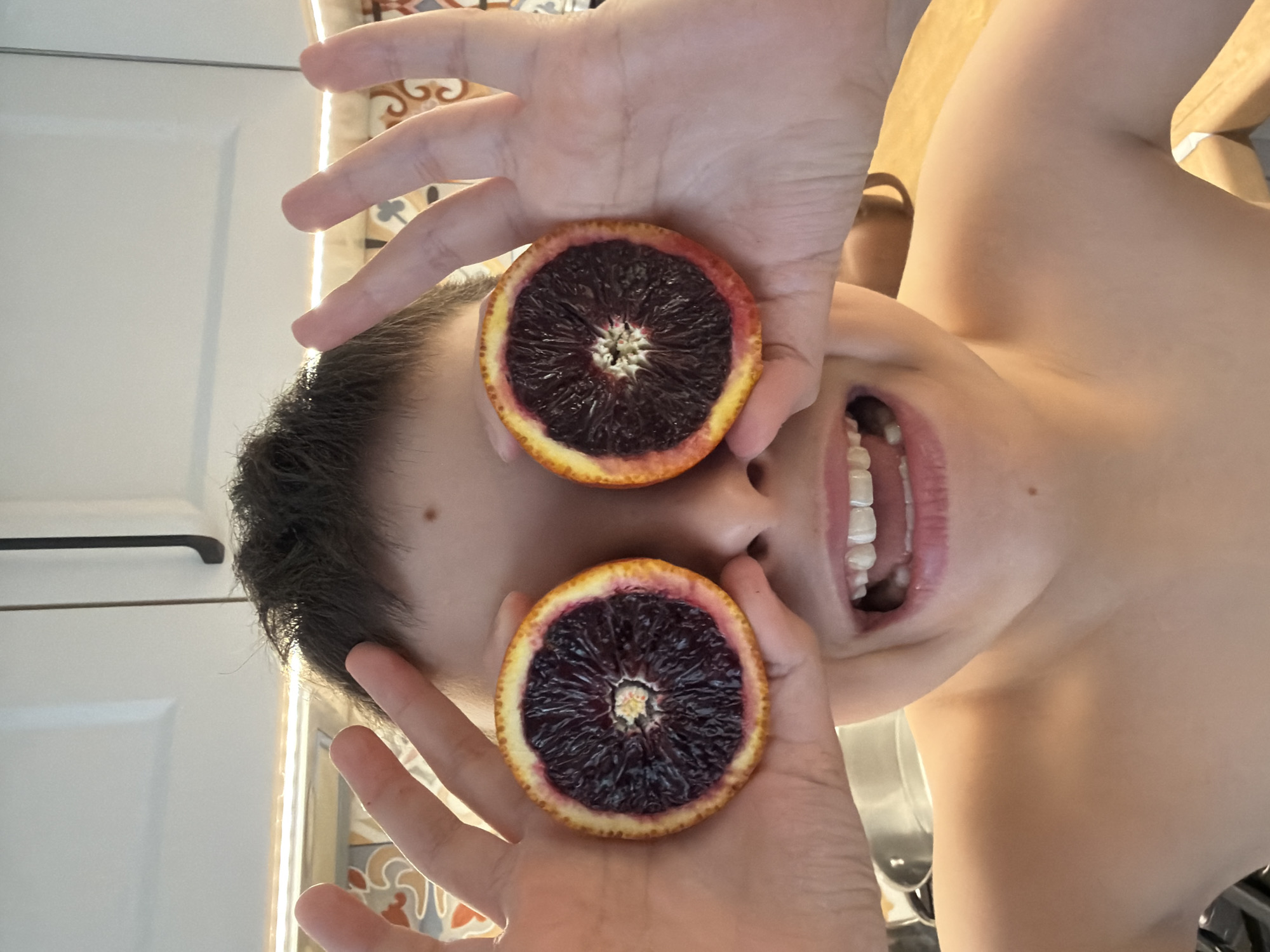 A close-up, slightly blurry, upside-down view shows a person holding two sliced blood oranges in front of their eyes. The person has short, dark hair and fair skin. Their mouth is open, revealing teeth. The background includes a white cabinet with a black handle and a strip of light. The image appears to be taken indoors. There is no indication of the location being Votkinsk, Russia, nor any other specific location.