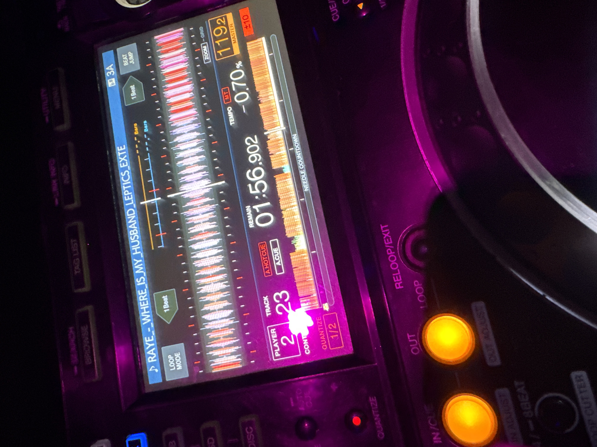The image displays a close-up, angled view of a professional DJ multi-player's illuminated screen and controls, set within a dimly lit environment in Moscow, Russia. The screen dominates the upper central portion, featuring a digital waveform representation of an audio track titled "RAYE_-_WHERE_IS_MY_HUSBAND_LEPTICS_EXTE". The track currently indicates "REMAIN 01:56.902" with a tempo adjustment of "-0.70%" and a master BPM of "119.2". The display also shows "PLAYER 2" and "TRACK 23", alongside "QUANTIZE 1/2" and "NEEDLE COUNTDOWN". Various function buttons are visible on the screen, including "LOOP MODE", "1 Beat", "BEAT JUMP", "SEARCH", "BROWSE", "TAG LIST", "LINK INFO", and "UTILITY MENU". Below the screen, physical controls are illuminated with a magenta hue, characteristic of a nightlife venue. Two prominent circular buttons, labeled "IN/CUE" and "OUT", glow bright orange. A red "QUANTIZE" button is visible to the lower left. A segment of a large, silver-rimmed jog wheel, illuminated with a soft white light, is partially visible in the lower right quadrant. No individuals are present in the frame.