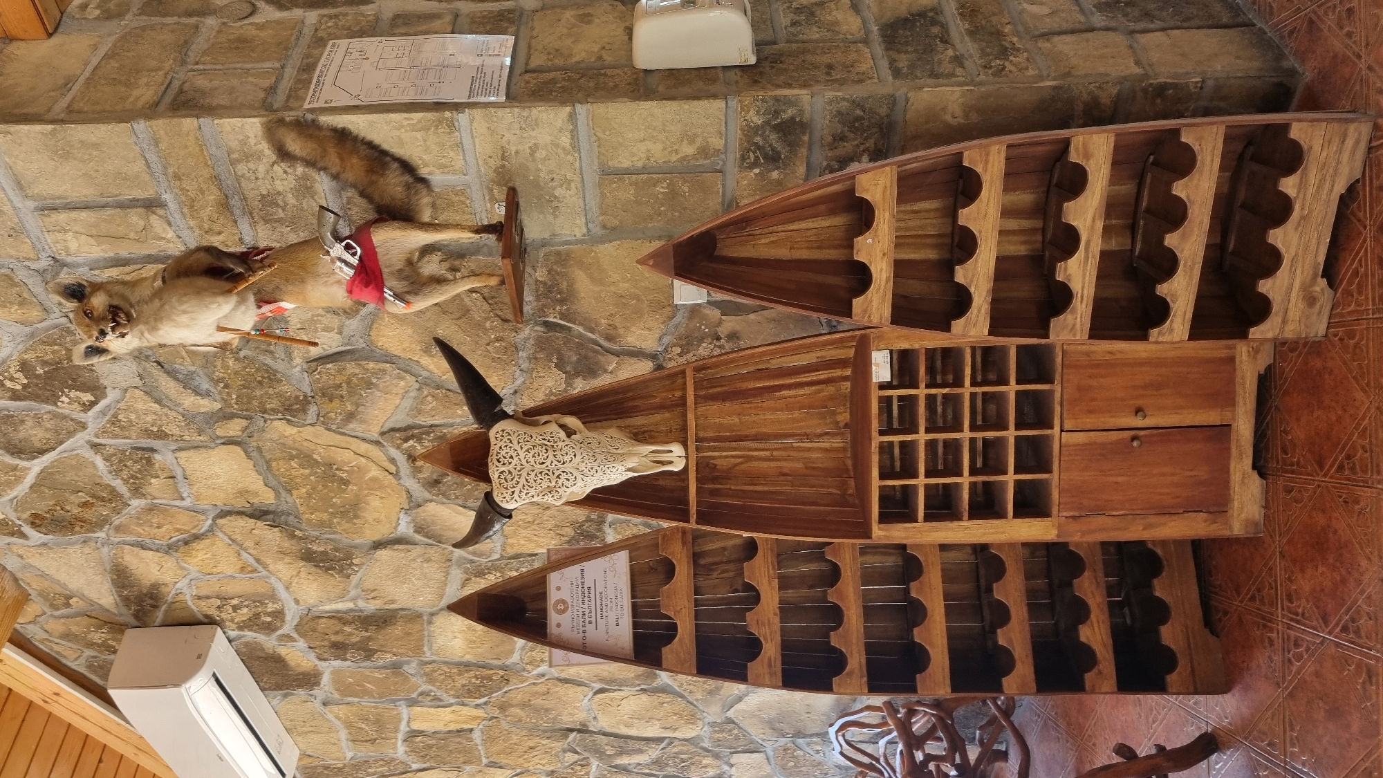 The image depicts a rustic interior with stone walls, wooden ceiling beams, and tiled flooring. Prominently featured are three tall, boat-shaped wooden shelves, likely for display or storage. The central shelf holds a decorative, carved skull with horns at the top. Above these shelves, mounted on the stone wall, is a taxidermied fox or coyote dressed in a red sash and holding two wooden sticks. A silver toy gun is attached to its sash. To the left of the shelves, a white air conditioning unit is mounted high on the wall. A printed map or diagram is visible on the wall to the right of the taxidermied animal. The overall aesthetic suggests a theme of a hunting lodge or a themed establishment. While the image is located in Zlatna Panega, Bulgaria, this is not visually evident from the image itself. There are no people or animals other than the taxidermied fox in the scene.