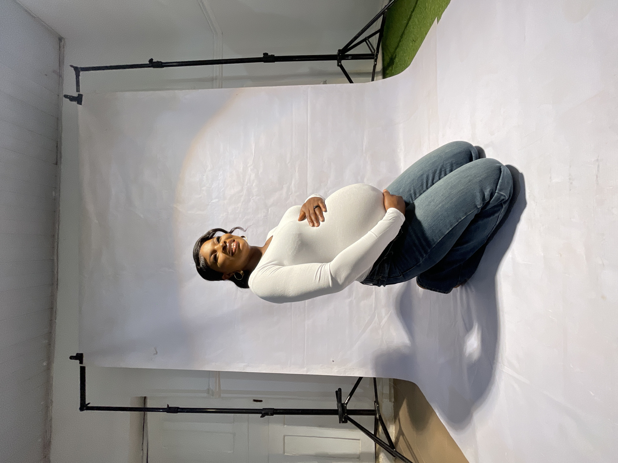 A pregnant woman lies curled on her side on a white backdrop. She is wearing a white, long-sleeved shirt and blue jeans. Her hands rest on her pregnant belly, and she is smiling. The backdrop is draped over a support structure consisting of black metal pipes. A white door is visible in the background on the lower left. Green artificial turf is visible in the upper right. The image was taken at an unspecified location. No information is available to confirm its location in the city of Jos, Nigeria.