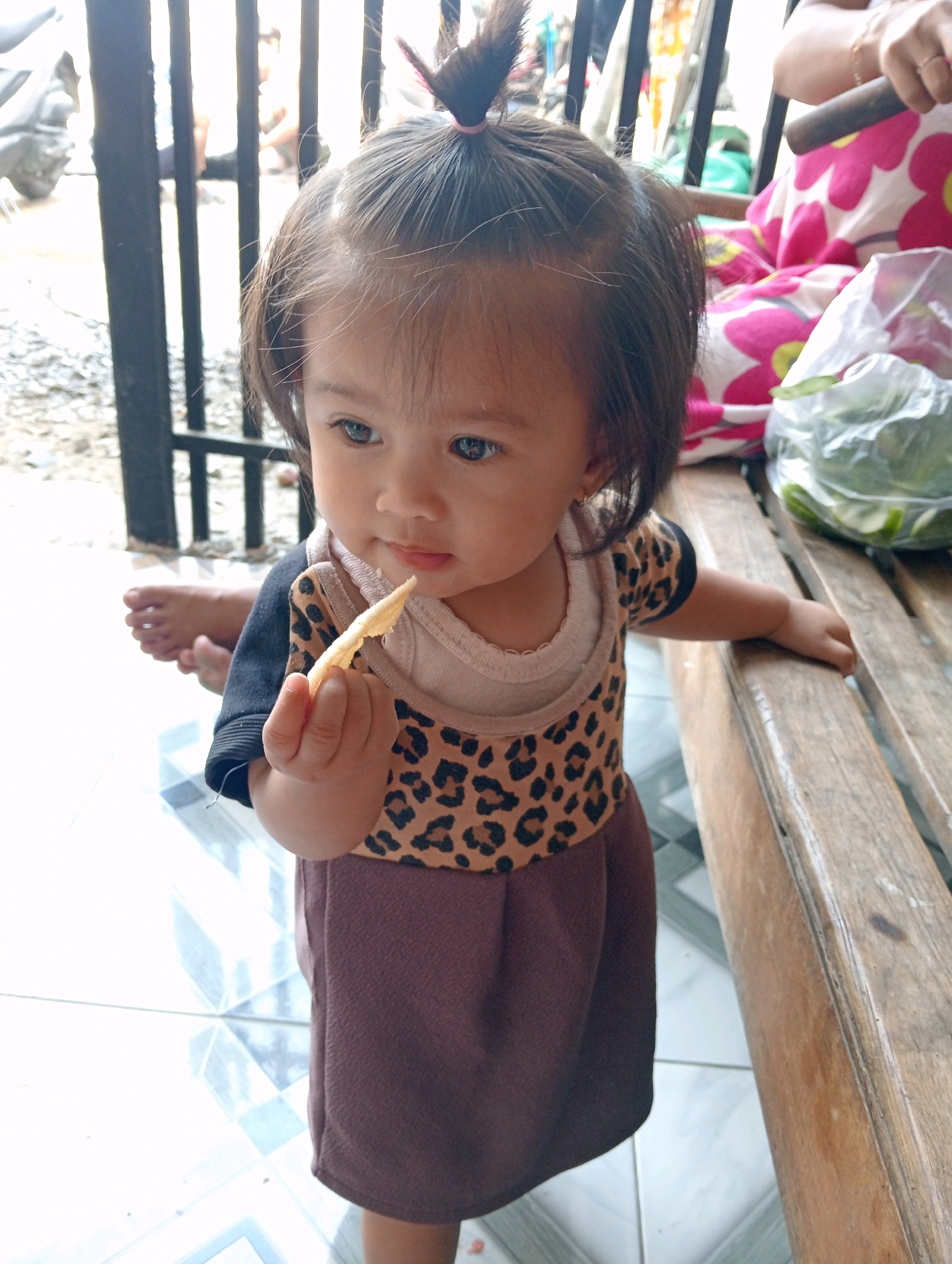 A young East Asian child, likely a toddler, stands in front of a black metal gate. The child has dark hair pulled into a small ponytail with a pink tie and is wearing a leopard-print t-shirt layered over a beige shirt, and a dark brown skirt. The child is holding a small, light-colored snack in their right hand and is looking down at it. In the background, through the gate, there are blurred figures and a motorcycle. To the child's right, a wooden bench is visible. Behind the child, a person wearing a floral patterned garment is seated, with a clear plastic bag containing green vegetables on the bench next to them. The ground beneath the child appears to be tiled. The image is taken from a slightly low angle, focusing on the child. The location is identified as Soko, Indonesia.