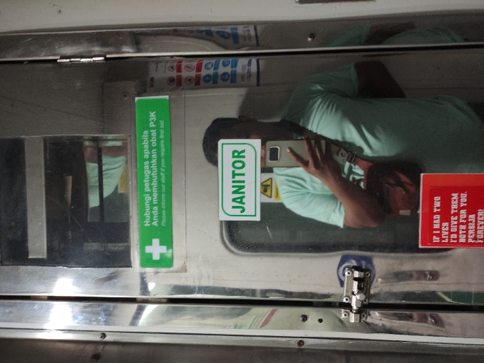 An image, rotated 90 degrees clockwise, captures a person's reflection in a shiny metal surface, likely a door or panel inside a train or similar public transport vehicle. The person, who appears to be taking the photo, is wearing a light green t-shirt, dark shorts, and a brown shoulder bag, and holds a smartphone to their face, obscuring it.
Several stickers are affixed to the reflective surface. A green rectangular sticker on the left, oriented vertically, displays text in Indonesian and English: "Hubungi petugas apabila Anda membutuhkan obat P3K" and "Please ask our staff if you require first aid," accompanied by a white cross symbol. Another green sticker, positioned centrally, reads "JANITOR" and includes a yellow warning triangle icon. To the right, a red sticker features the text: "IF I HAD TWO LIVES I'D GIVE THEM BOTH FOR YOU. PERSIJA FOREVER!"
Other details visible include a metal hinge or latch mechanism at the bottom center of the frame and faint reflections of interior elements. The setting is indoors, with artificial lighting. The combination of Indonesian text and the "JANITOR" sign suggests a public service area. The "PERSIJA" text on the red sticker refers to an Indonesian football club. The scene is located in Purwosari, Indonesia.