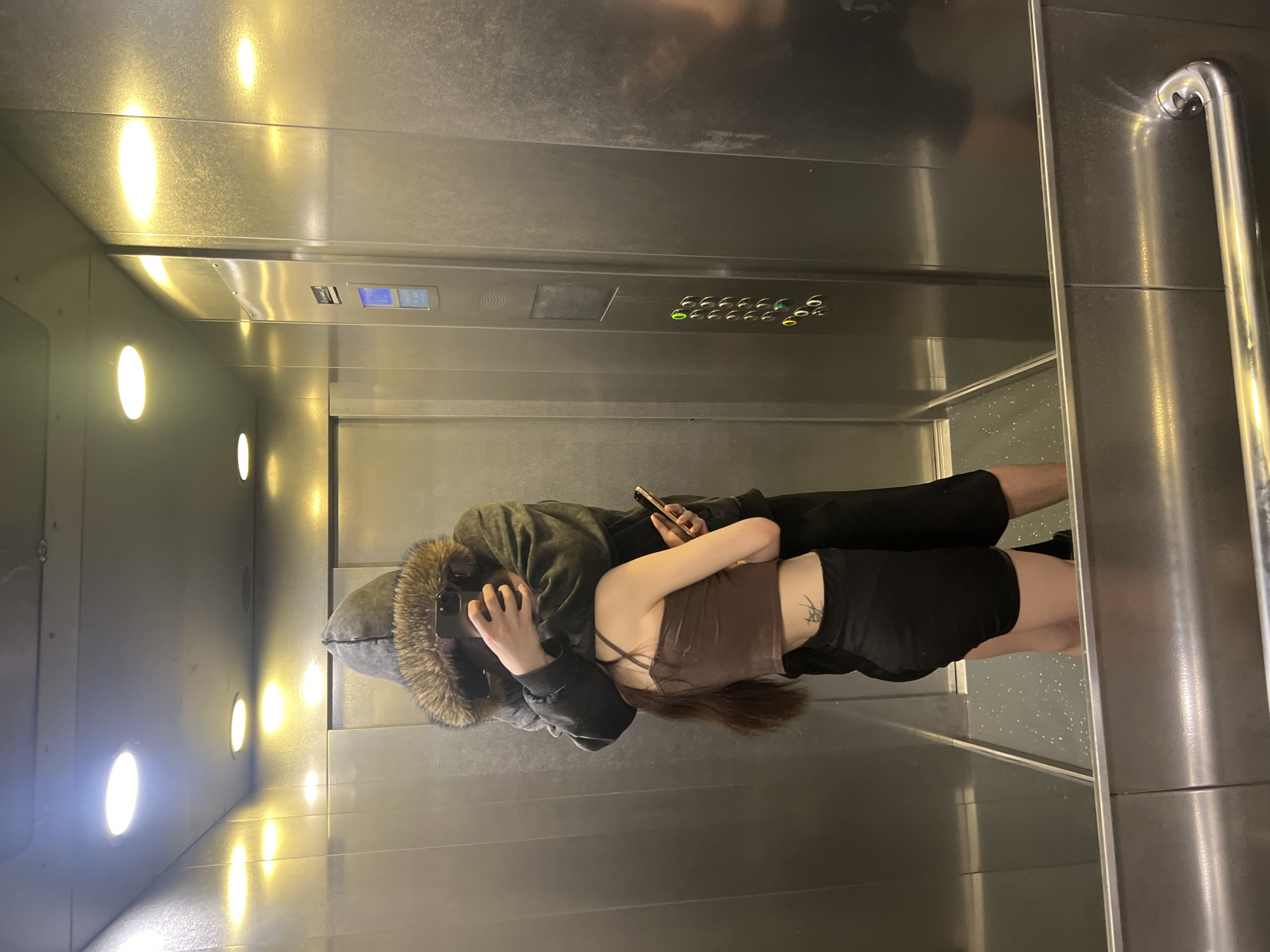 The scene is set inside a modern reflective elevator in Saint Petersburg, Russia. Two individuals are depicted in an intimate embrace. The taller individual, positioned closer to the camera, wears a dark grey hooded jacket with a prominent fur trim, obscuring their face with a black smartphone held in their right hand. Their reflection shows their full height, clad in dark trousers. The shorter individual, embraced by the first, wears a brown crop top and black shorts, exposing their midriff and a dark tattoo on their left lower back/side. Their long dark hair hangs down their back. They hold a black smartphone in their left hand, visible to the side.
The elevator interior features polished metallic walls and ceiling, highly reflective, mirroring the occupants and internal lights. Recessed ceiling lights illuminate the space; some are bright white, while others emit a warmer, yellow glow, casting reflections and highlights across the metal surfaces. The right wall features a control panel with a blue digital display and a vertical column of circular buttons, some illuminated green and yellow, indicating floor selections. The floor has a speckled, possibly dark grey or black, surface with white flecks. A chrome handrail is visible in the lower right reflection. The metallic walls show minor scuffs and texture variations.