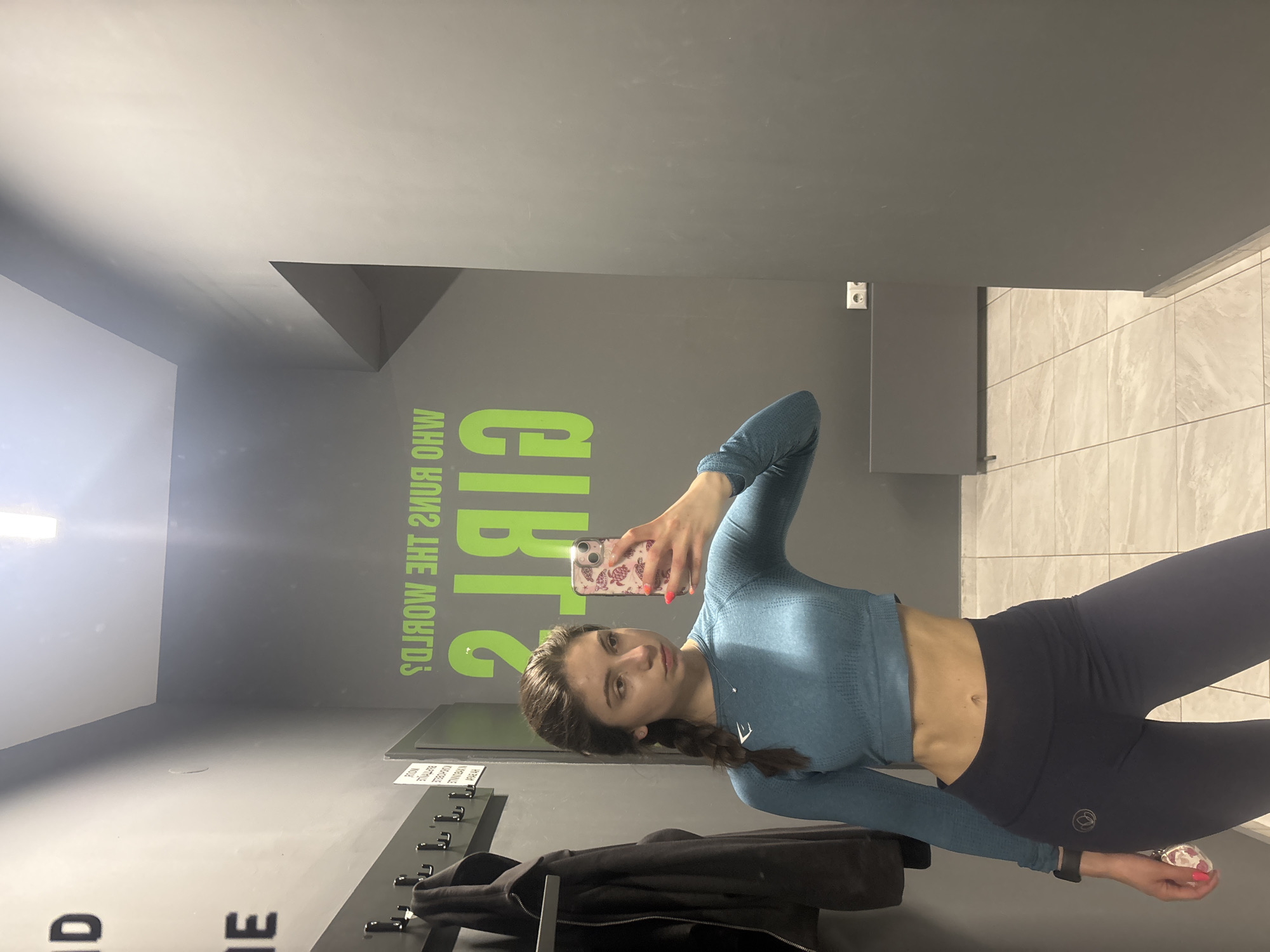 A young woman is captured in a mirror selfie, standing indoors in what appears to be a gym changing room or facility in Varna, Bulgaria. The image is oriented sideways, showing the subject in athletic wear.
The woman has dark hair styled in a braid and is wearing a long-sleeved, teal-colored textured crop top that reveals her midriff, and dark blue or black leggings. A black watch is visible on her left wrist, and a small white logo, possibly an 'E' or similar design, is present on the front of her top. A subtle circular logo can also be seen on her leggings. She holds a smartphone with a patterned case to take the picture, her fingernails painted bright pink. Her expression is neutral and confident.
The background features dark gray walls. On the left wall, a prominent green graphic spells out "GIRLS WHO RUNS THE WORLD?" (reversed due to the mirror reflection, indicating the actual text is "WHO RUNS THE WORLD? GIRLS"). Below this text, a coat rack with multiple hooks holds a black jacket. A partially visible, reversed sign with text is also present near the coat rack. To the right of the woman, the wall transitions into light-colored tiles, possibly indicating a shower area or another section of the facility. An electrical outlet is visible on the upper right side of the dark gray wall. Artificial lighting illuminates the space.