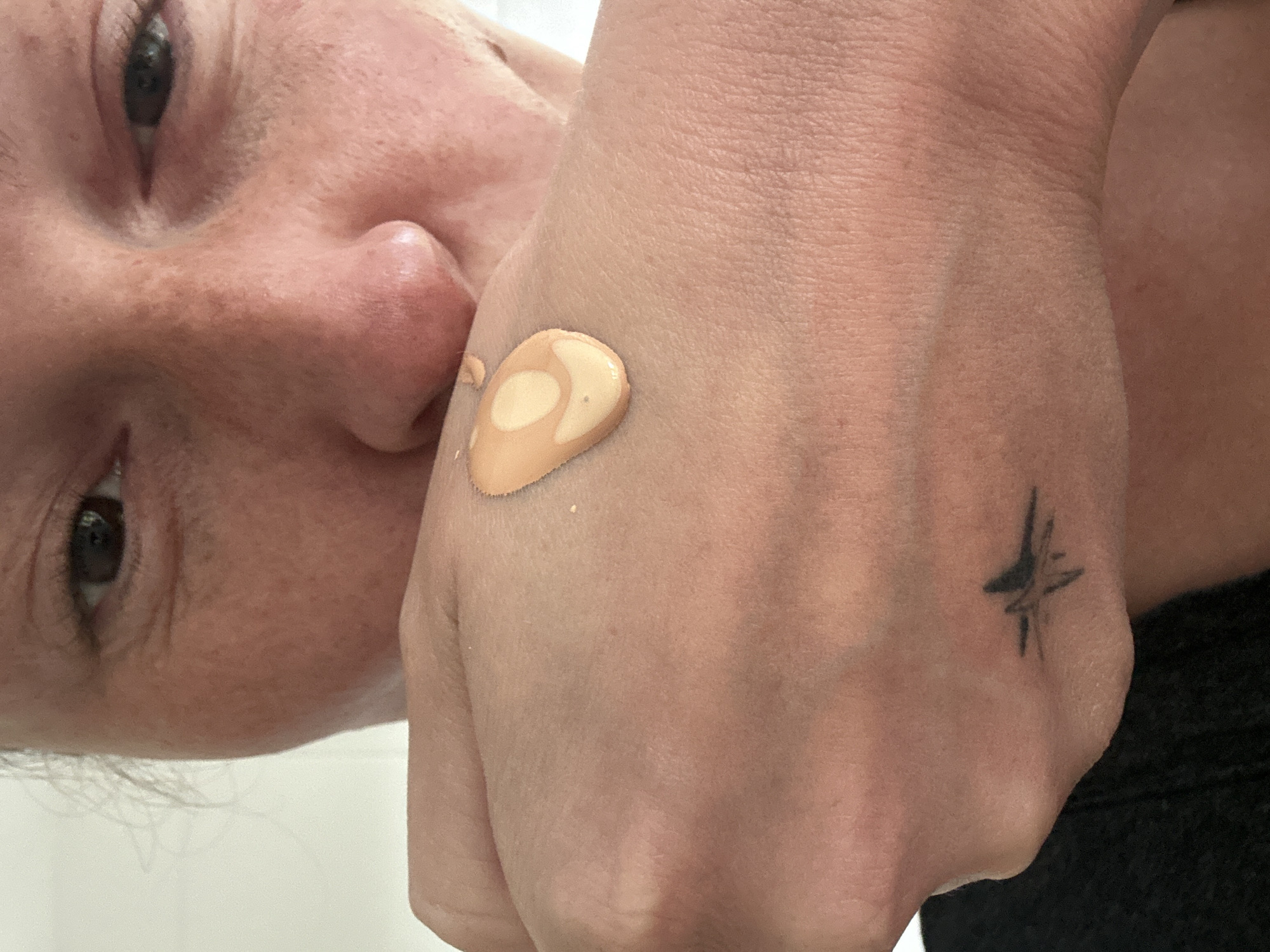 A close-up, tilted shot depicts a person with fair skin, testing a drop of beige-toned foundation on the back of their hand. The person's face, including their eyes, nose, and cheeks, is visible in the frame. A small tattoo of a star-like symbol can be seen on the wrist of the hand holding the foundation. The background appears to be a plain white or light-colored surface. The location is not identifiable from the provided image.