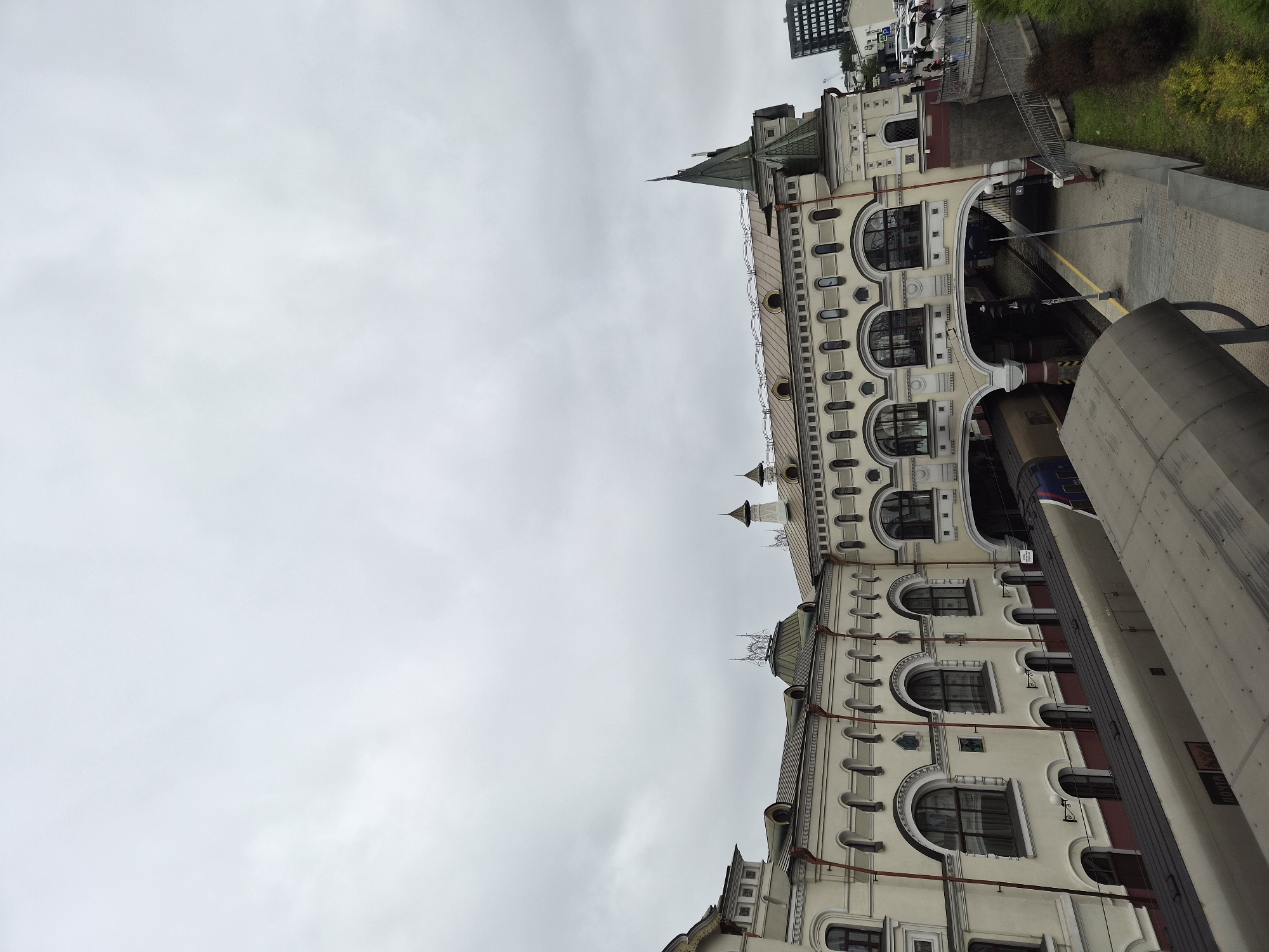The image shows a view of the Vladivostok railway station in Vladivostok, Russia, on an overcast day. The station building, with its distinctive architectural style, is prominently featured. A train is parked at the platform, partially visible in the foreground. The sky is filled with clouds, casting a muted light on the scene. The platform is paved, and there is a grassy embankment to the right. A few people are visible on the platform and on a walkway leading up to the station. A modern, dark-colored building is visible in the background to the right, contrasting with the historic architecture of the station. The scene appears calm, with no apparent activity or interaction of note.