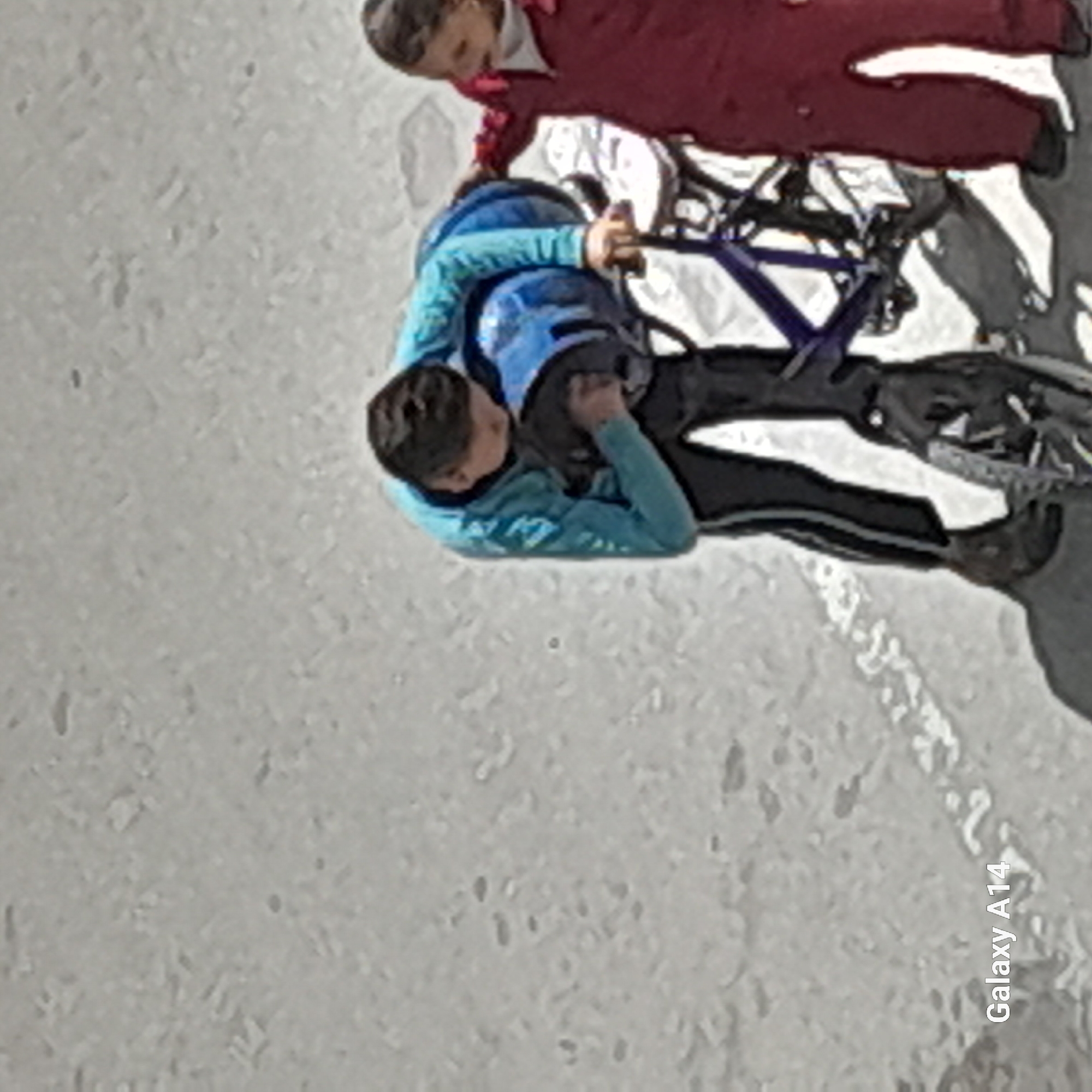 Here is a forensic description of the image:
Two individuals are present, positioned on a bicycle. One individual, dressed in red attire, sits in front. The other, wearing blue, is behind, in what appears to be in the position of control. The location is in a city. The immediate surrounding is concrete. The image contains an unidentified text in the bottom right corner.