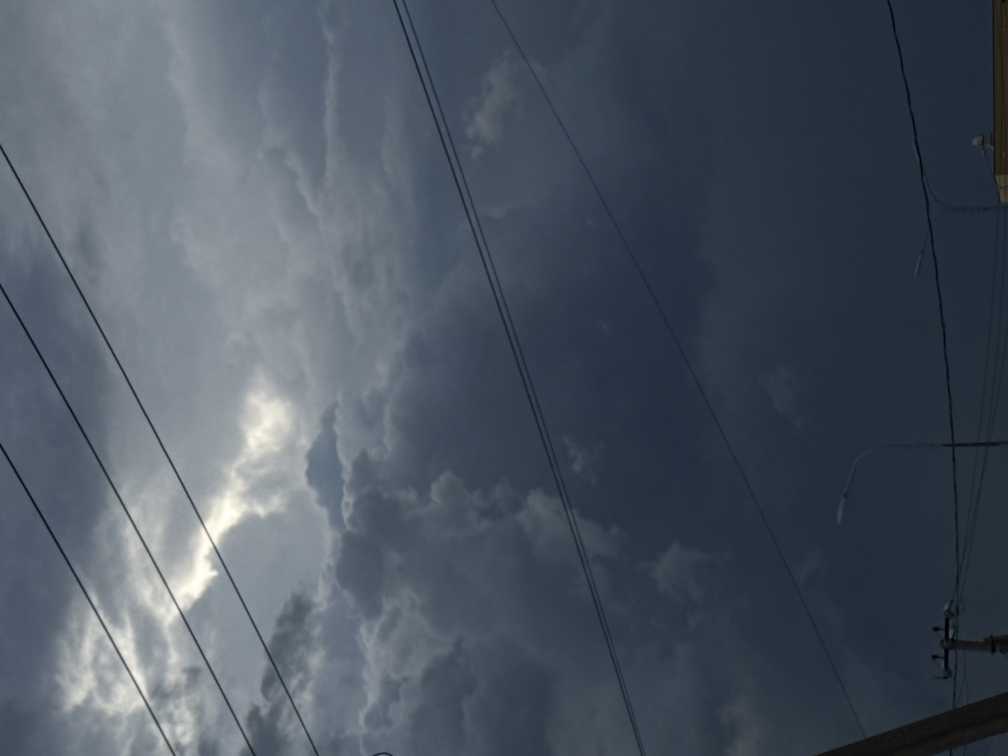 The image depicts a sky filled with dark and dramatic storm clouds. Power lines run diagonally across the frame from the left, intersecting with the clouds. A utility pole with a street lamp is partially visible in the lower right corner. The contrast between the dark clouds and a patch of brighter light creates a sense of depth and impending weather. The location is Unknown, Unknown. No people or subjects are visible. Actions include the presence of atmospheric activity and the infrastructure of the utility pole.