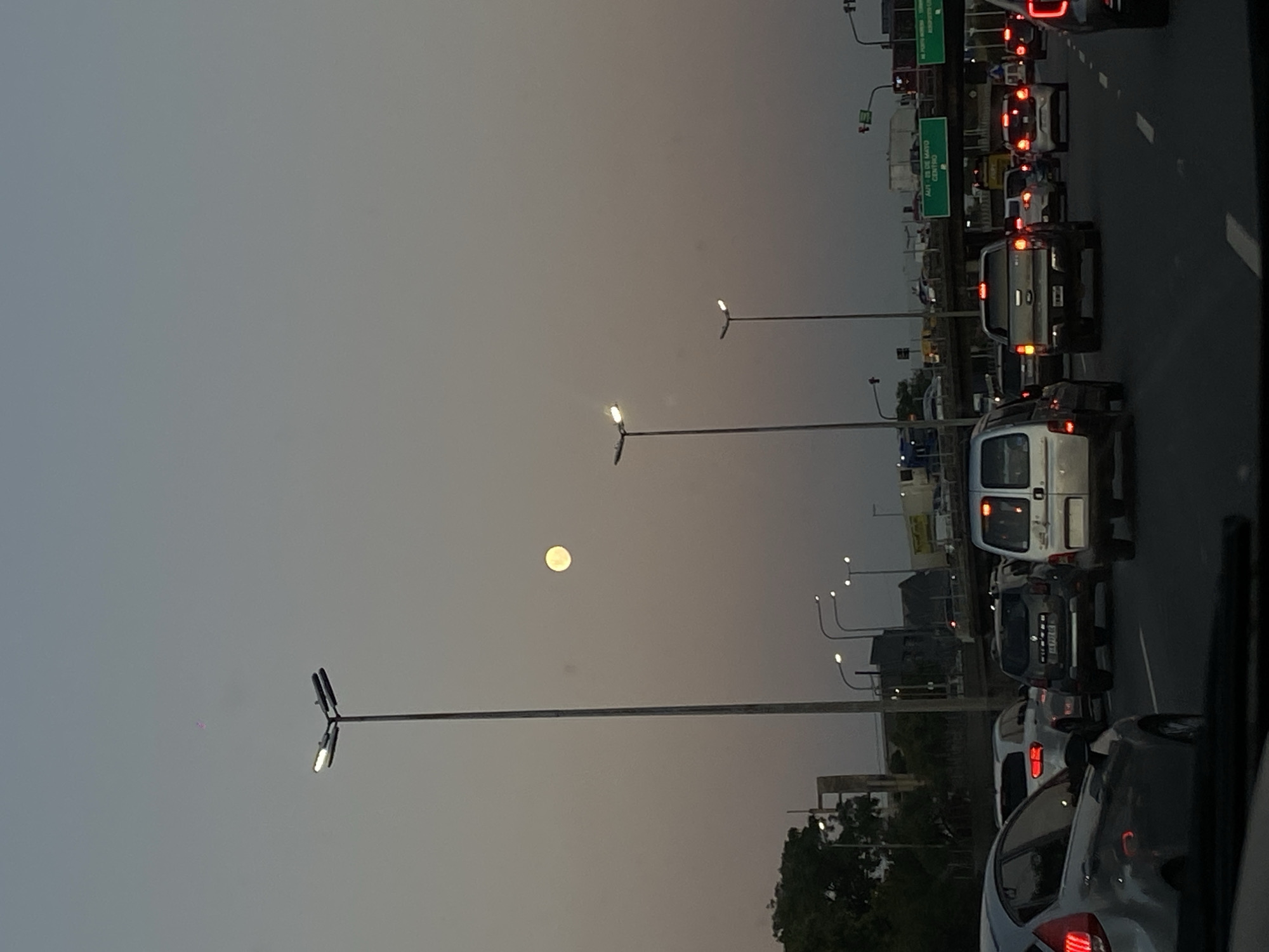 The image depicts a traffic jam on a highway in Buenos Aires, Argentina, at dusk. The view is from inside a car, facing forward and slightly to the right. The sky is a hazy gray-brown. In the sky, a bright full moon is visible. Overhead streetlights line the highway. On the right are lanes of stationary or slow-moving vehicles. Some of the car lights are on. A sign that says "AUT. 25 DE MAYO CENTRO" is visible. Trees line the side of the highway.