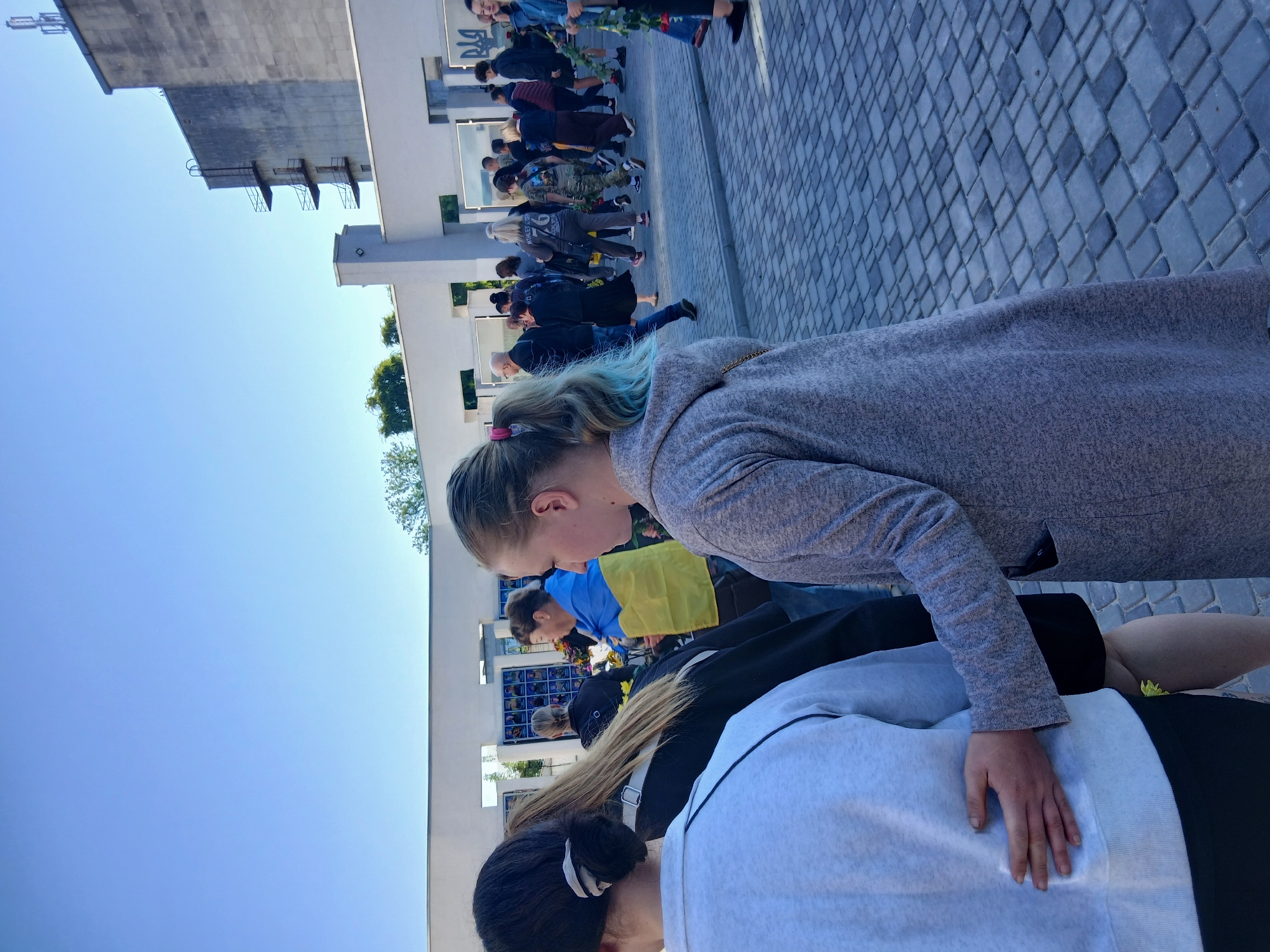 This is a full, eye-level shot of a group of people walking on a paved area outside. In the foreground, a young woman with light blue-tipped hair is looking to the right, wearing a gray hooded jacket. A hand is resting on her back. Behind her, a woman wearing a black jacket and another woman wearing a Ukrainian flag draped over her shoulders are walking. In the background, a large, Brutalist-style building with balconies and a water tower is visible. The ground is paved with gray interlocking bricks. A wall with what appears to be a memorial with photos is visible in the middle ground. The sky is clear and blue. The scene appears to be a public space in Verkhnodniprovsk, Ukraine, likely a memorial or solemn gathering.