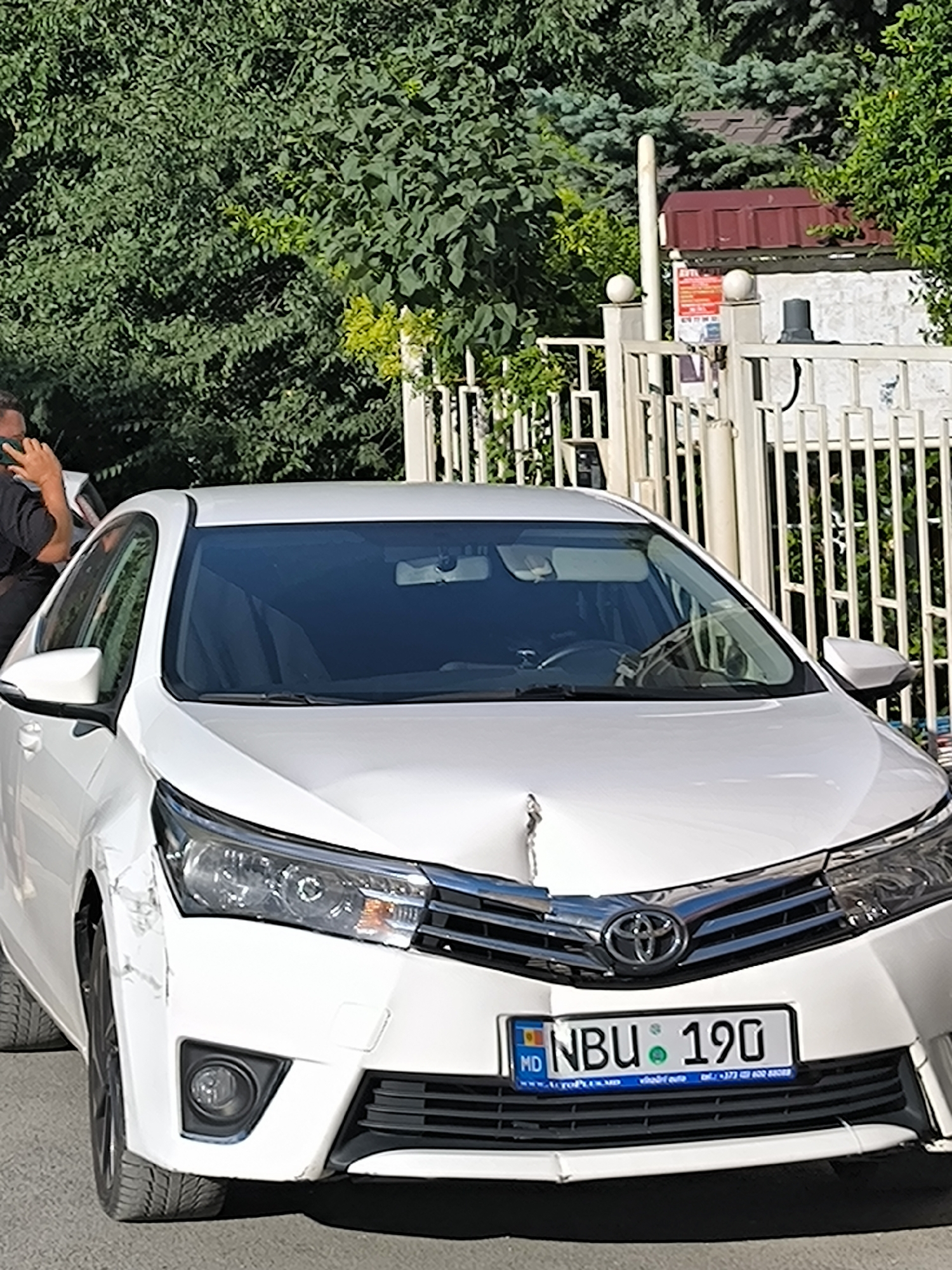 A white Toyota Corolla with visible front-end damage, including a dented hood and scraped bumper, is parked on a paved surface. The car has a Moldovan license plate that reads "NBU 190". In the background, a white metal fence with vertical slats is visible, along with lush green trees and a building with a reddish-brown roof. A person, partially visible on the left, is holding a phone to their ear. The scene appears to be outdoors in an urban setting, likely in Chișinău, Moldova, given the license plate and the architectural style of the building. The lighting suggests it is daytime.