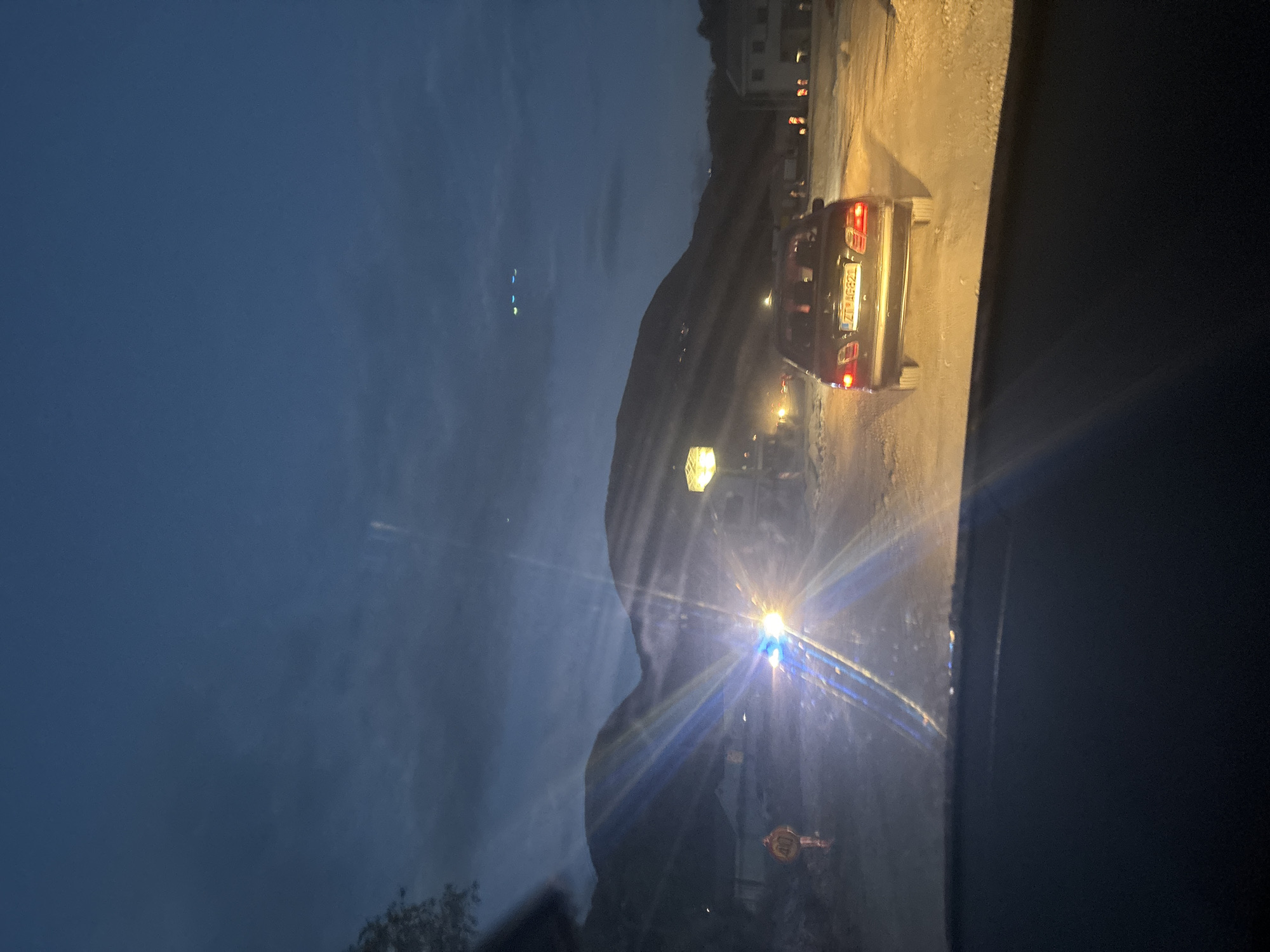 The image depicts a night scene in Lastva Grbaljska, Montenegro, viewed from inside a vehicle. Another car with the license plate "ZT AG621" is directly ahead, its tail lights illuminated. Streetlights and building lights are visible in the background, along with a dark mountain silhouette. The sky is a dark blue, suggesting dusk or night.