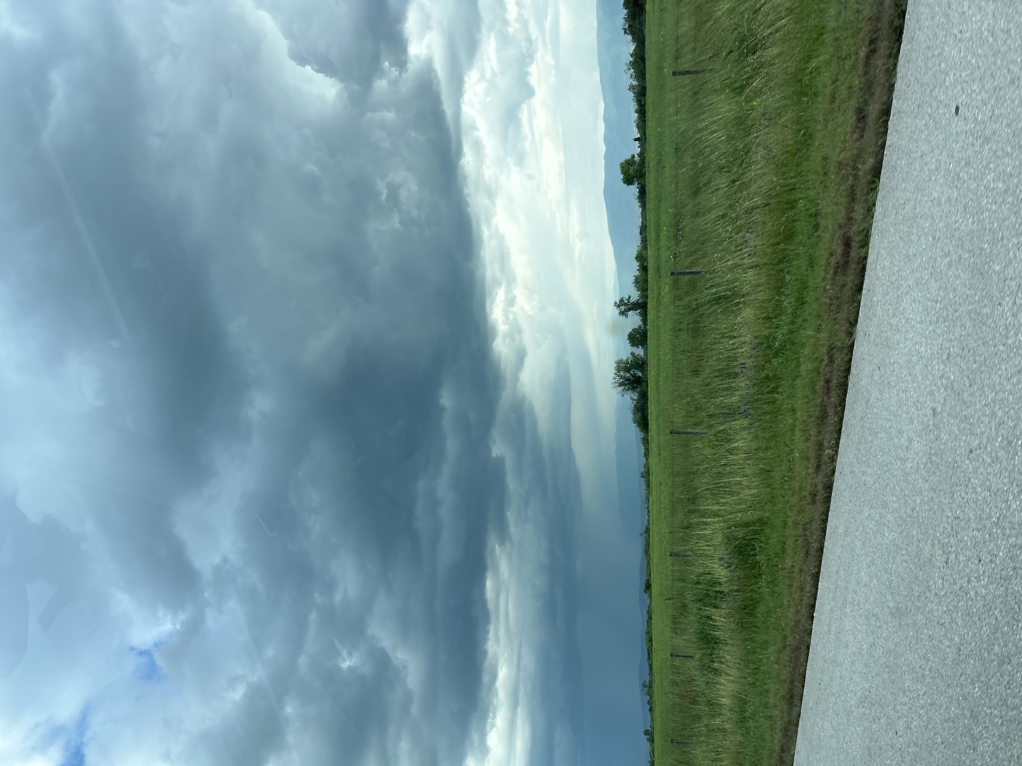 The image depicts a landscape view, likely taken from a moving vehicle. The sky is filled with dark, stormy clouds, suggesting an approaching weather event. A strip of green grassland is visible on the right side of the image, marked by occasional trees and wooden fence posts. In the distance, faint outlines of mountains can be discerned beneath the cloud cover. A portion of a grey road surface is visible in the lower right corner, further indicating the image was captured from a vehicle on a road. There is no information in the image that places it in Shtarkovo, Bulgaria.