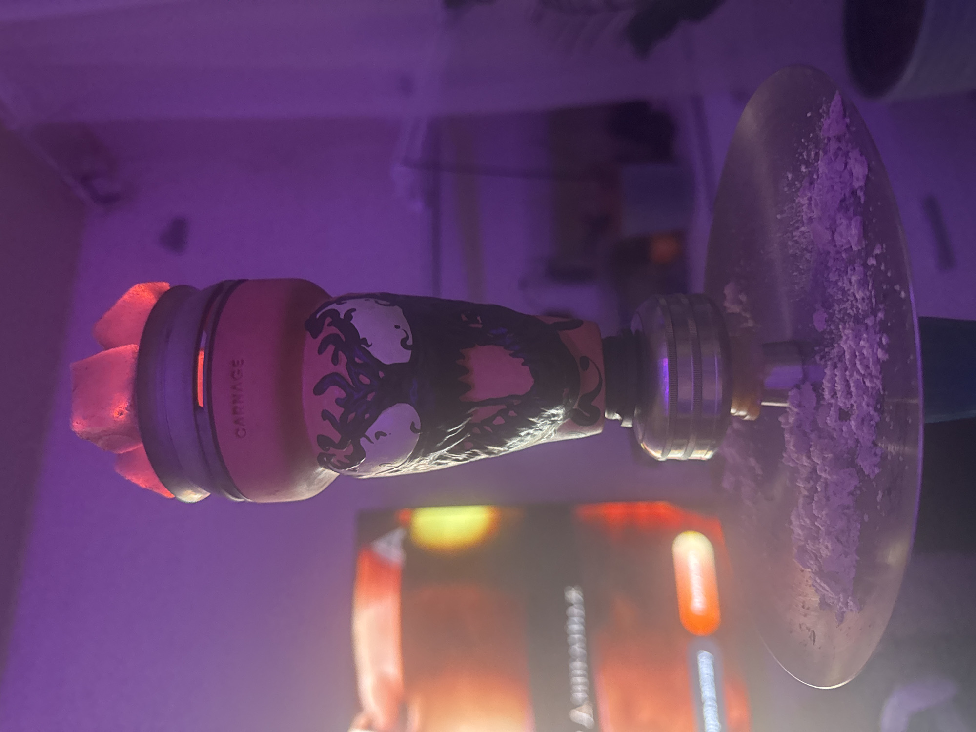 A close-up indoor shot depicts a hookah lying on its side, featuring a "Carnage" character design on its body. The hookah head holds lit coals. A metallic plate attached to the main stem is dusted with a white substance. The image is taken in an environment with purple ambient lighting. The location appears to be inside a room, possibly in Chaikovsky, Russia, but there's no specific evidence in the image to confirm the exact city. There are no people visible in the frame. The foreground is slightly blurred.
