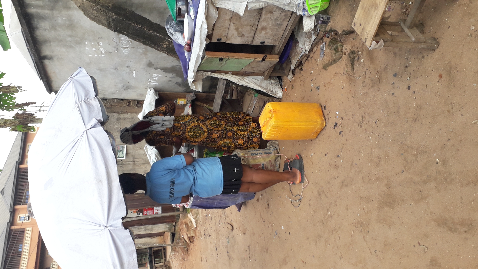 The image depicts an outdoor scene in Nkpor, Nigeria, likely a market or roadside stall. In the foreground, a person wearing a blue t-shirt with "YUUQO FAMILY" printed on the back and black shorts stands facing away from the camera. They are wearing dark sandals with straps. To the left of this person is a large, white umbrella, mostly obscuring another person and potentially merchandise beneath it.
Behind the person in the blue shirt, a woman in a vibrantly patterned, long dress with yellow and brown motifs is visible. She appears to be standing near some wooden structures that form part of a stall. A large, bright yellow plastic container is positioned on the sandy ground near the stall. The ground itself is covered in sand and scattered debris.
The setting appears to be rustic, with makeshift structures and a general sense of a busy, informal trading environment. The lighting suggests it is daytime. There are no explicit cues about the weather, but the overall scene is consistent with a warm climate. The people's posture doesn't convey strong emotions, but the scene suggests daily commercial activity. Visible text includes "YUUQO FAMILY" on the t-shirt and some unreadable text on bags and possibly on the stall structure.