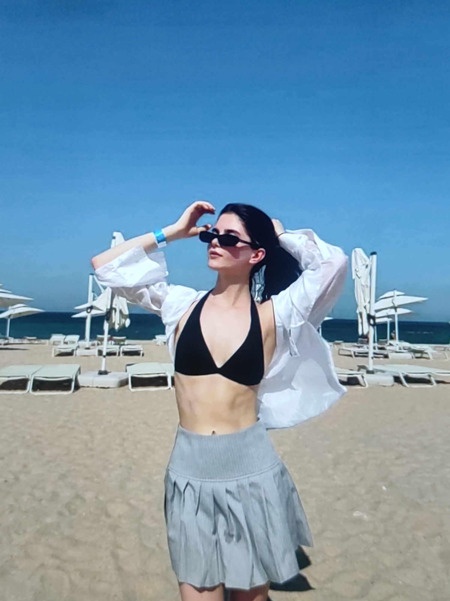 The image shows a young woman standing on a sandy beach. She is wearing a black bikini top, a light-colored, unbuttoned shirt, and a grey pleated skirt. She has on dark sunglasses and is holding her hands up, seemingly adjusting her hair. Her hair is dark and appears to be pulled back. In the background, there are lounge chairs and beach umbrellas, suggesting a resort or public beach area. The sky is clear and blue, indicating good weather, likely daytime given the brightness of the scene. The ocean is visible in the distance. There is no visible text in the image. The overall impression is one of leisure and enjoyment at a beach destination.