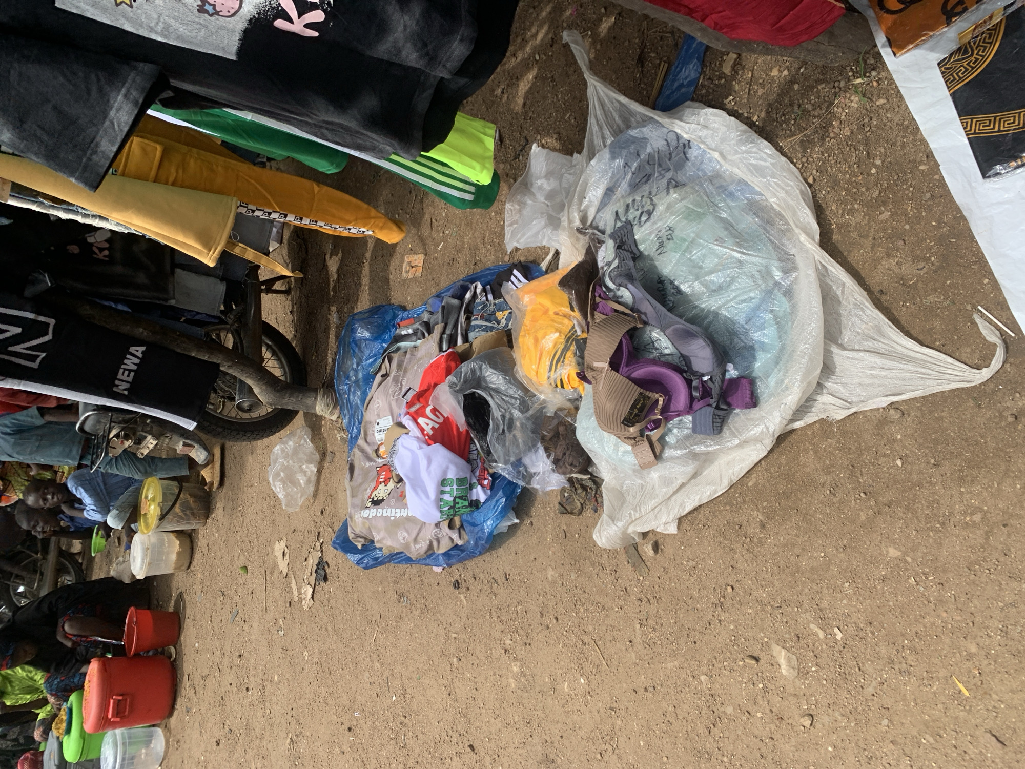 This is an outdoor scene in what appears to be a market in Project, Nigeria. The ground is dirt and appears dusty. In the foreground and midground, there are piles of clothing and other items. To the left, there are several large red and green plastic containers. Behind these containers, several people are seated on the ground. In the center and right of the image, there are items of clothing hanging from a rack or structure, including t-shirts and pants in various colors. Some of the clothing items are also piled on the ground, some wrapped in plastic. A motorcycle is visible behind the hanging clothes. The overall impression is of a busy, informal market setting.