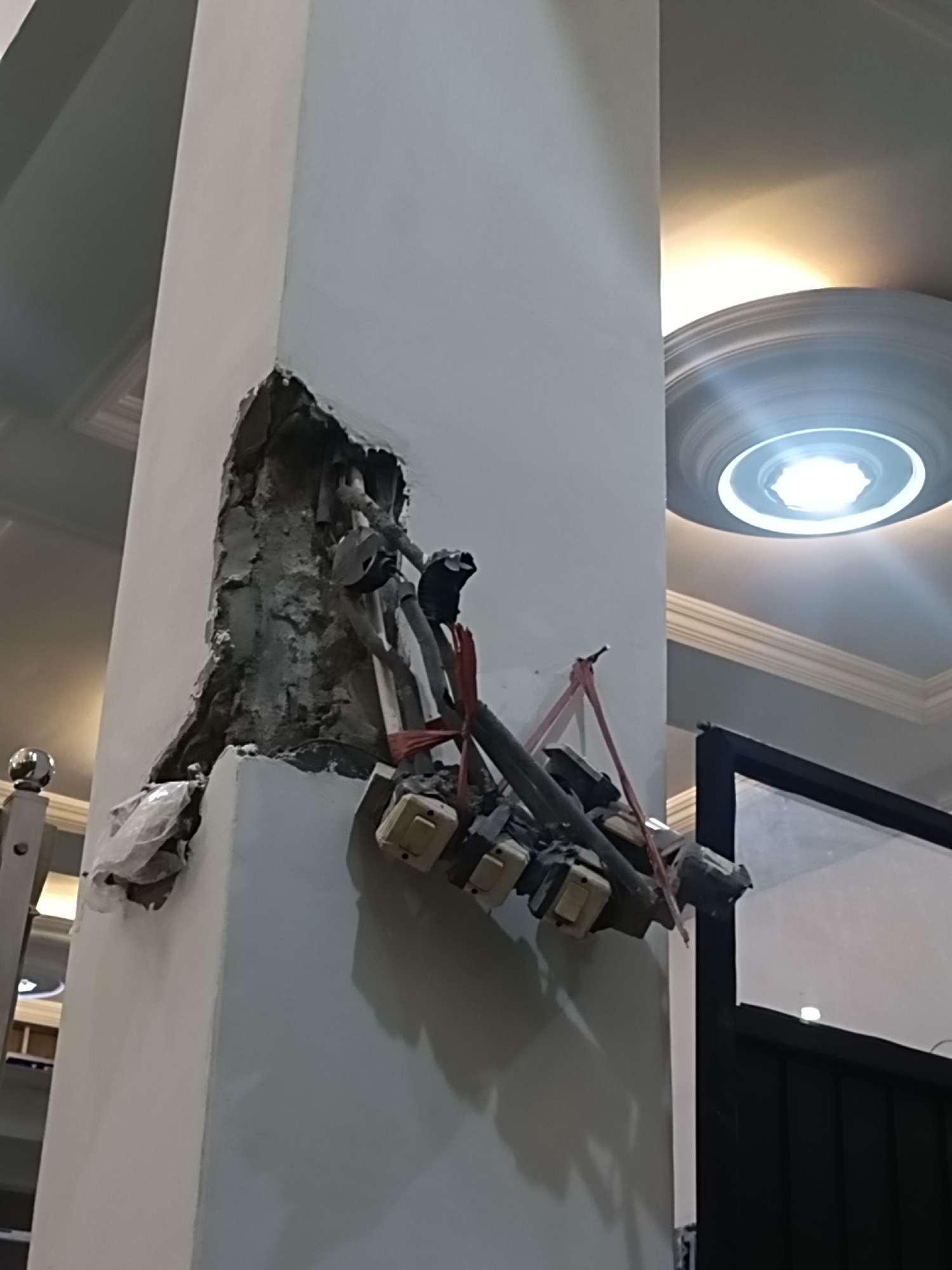 The image depicts a damaged concrete pillar with exposed electrical wiring and switches. The damage to the pillar appears to be a result of impact, with a large section of the concrete broken away, revealing the internal structure and conduits. Several electrical components, including switches and wiring, are haphazardly attached to the exposed structure and secured with red ties. The scene is indoors, suggested by the presence of a ceiling with decorative molding and a circular light fixture with a bright light. A portion of a dark framed object, possibly a door or window, is visible on the right side of the frame. The overall impression is one of disrepair and unfinished or damaged electrical work within a building.