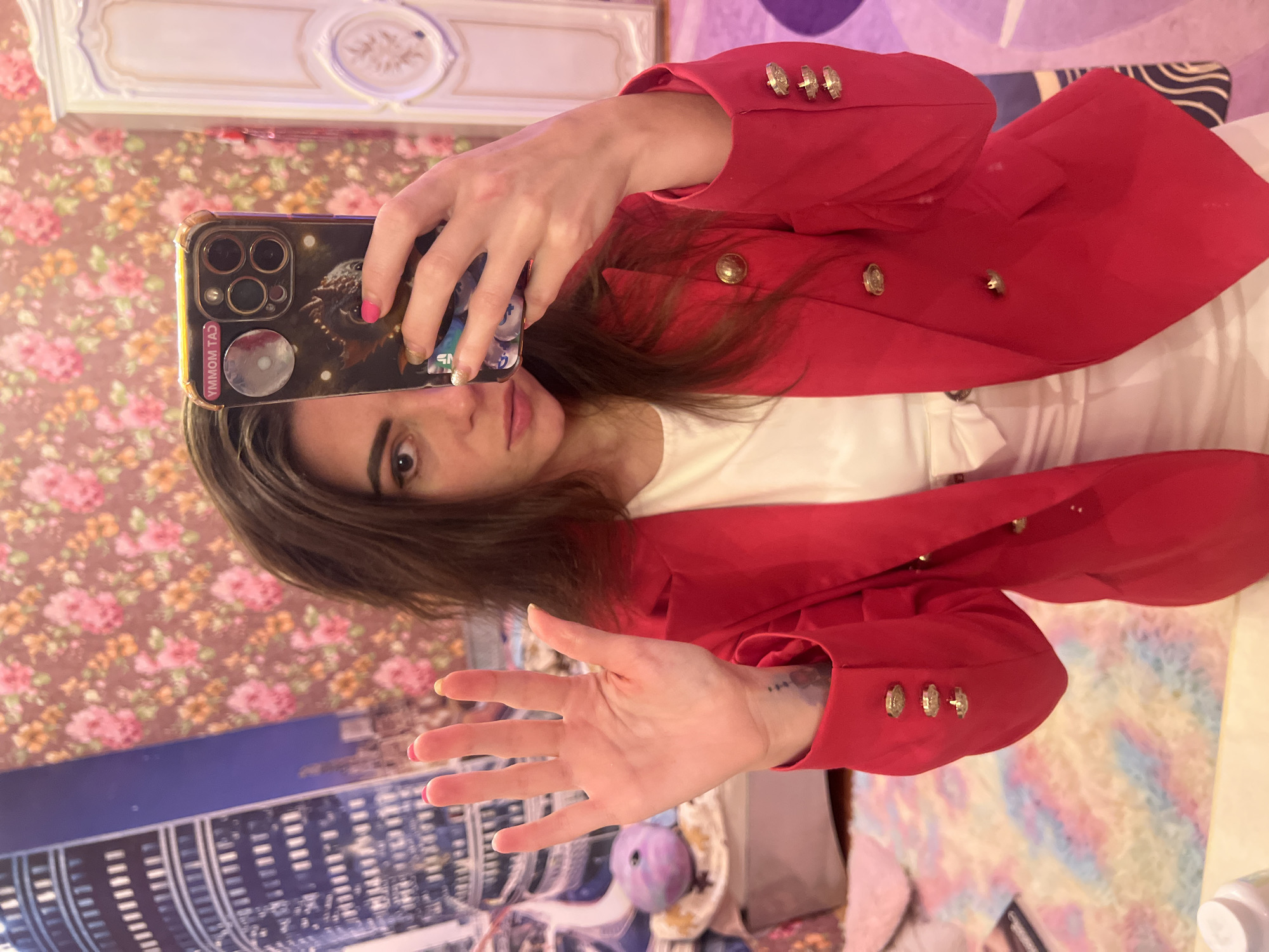 A woman is taking a selfie in a room with floral wallpaper. She is wearing a red blazer over a white top and is holding up her left hand, showing five fingers. Her right hand is holding a smartphone. In the background, there is a large tapestry of a cityscape. The room appears to be decorated in a feminine and possibly eclectic style, with a hint of vintage furniture. The woman has dark hair and is looking at the camera.