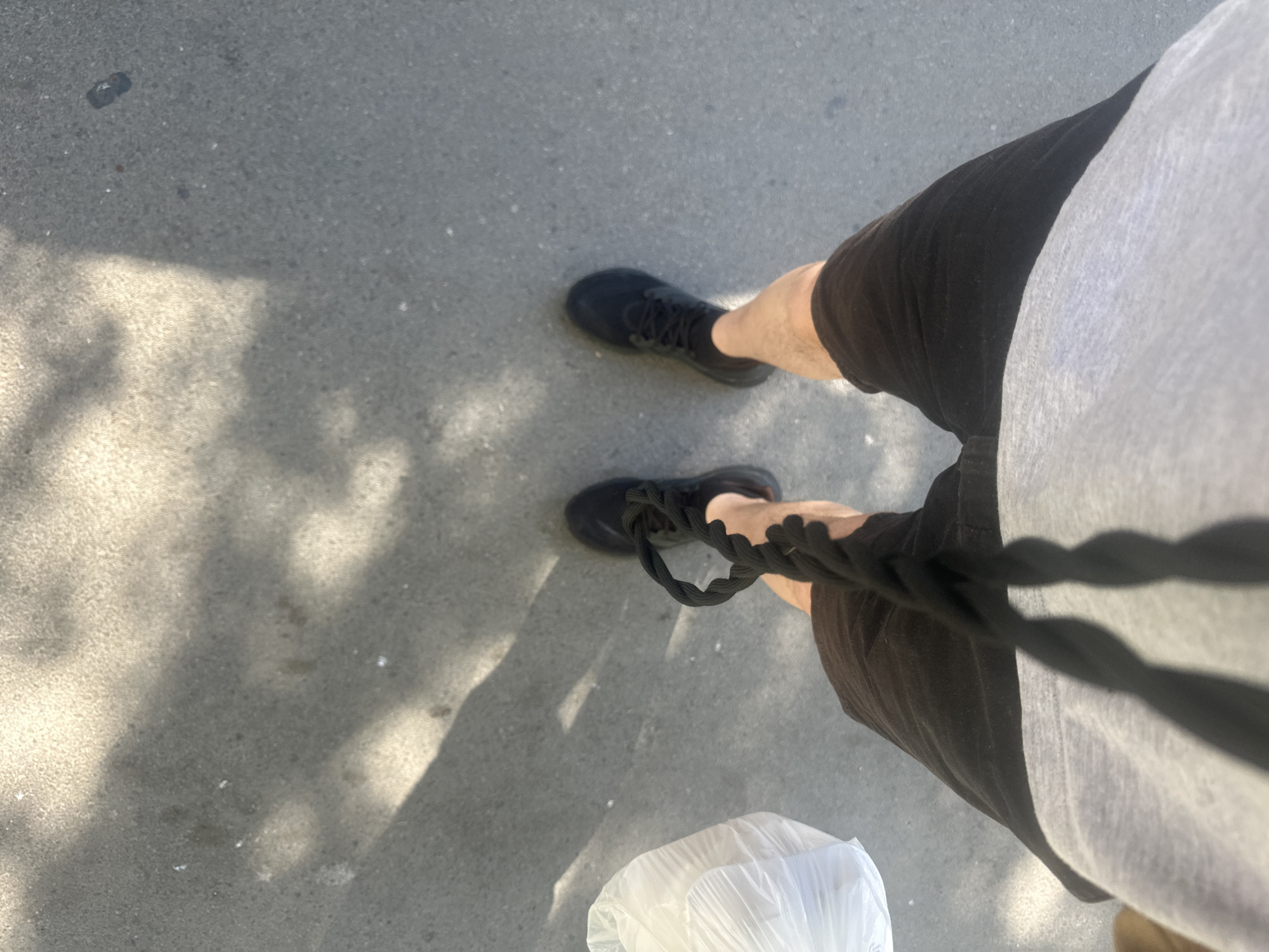 The image shows a person standing on a concrete surface, likely a sidewalk in an urban area. The person is wearing black sneakers, dark shorts, and a light-colored t-shirt. They are holding a black rope or cord, which casts a shadow across the frame. A white plastic bag is visible near their feet. The presence of shadows suggests it is daytime with sunlight. The location is not identifiable as Budva, Montenegro based on the visual information provided.