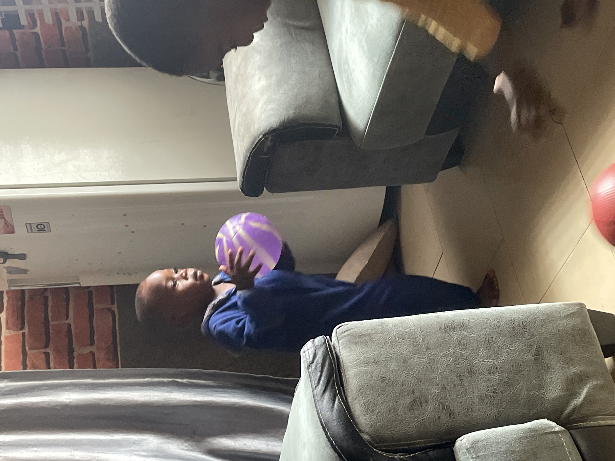 The image depicts an indoor scene, likely a living room, with two young children. On the left, a young boy in a blue outfit stands and holds a purple and white striped ball. He is positioned between a large, textured gray sofa and a silver-colored appliance, possibly a refrigerator. The wall behind him features a brick pattern. To the right, another child, seen from the back and in silhouette, is partially visible, facing the sofa. The floor is tiled. The context of Jos, Nigeria, is not discernible from the visual elements of the photograph itself.